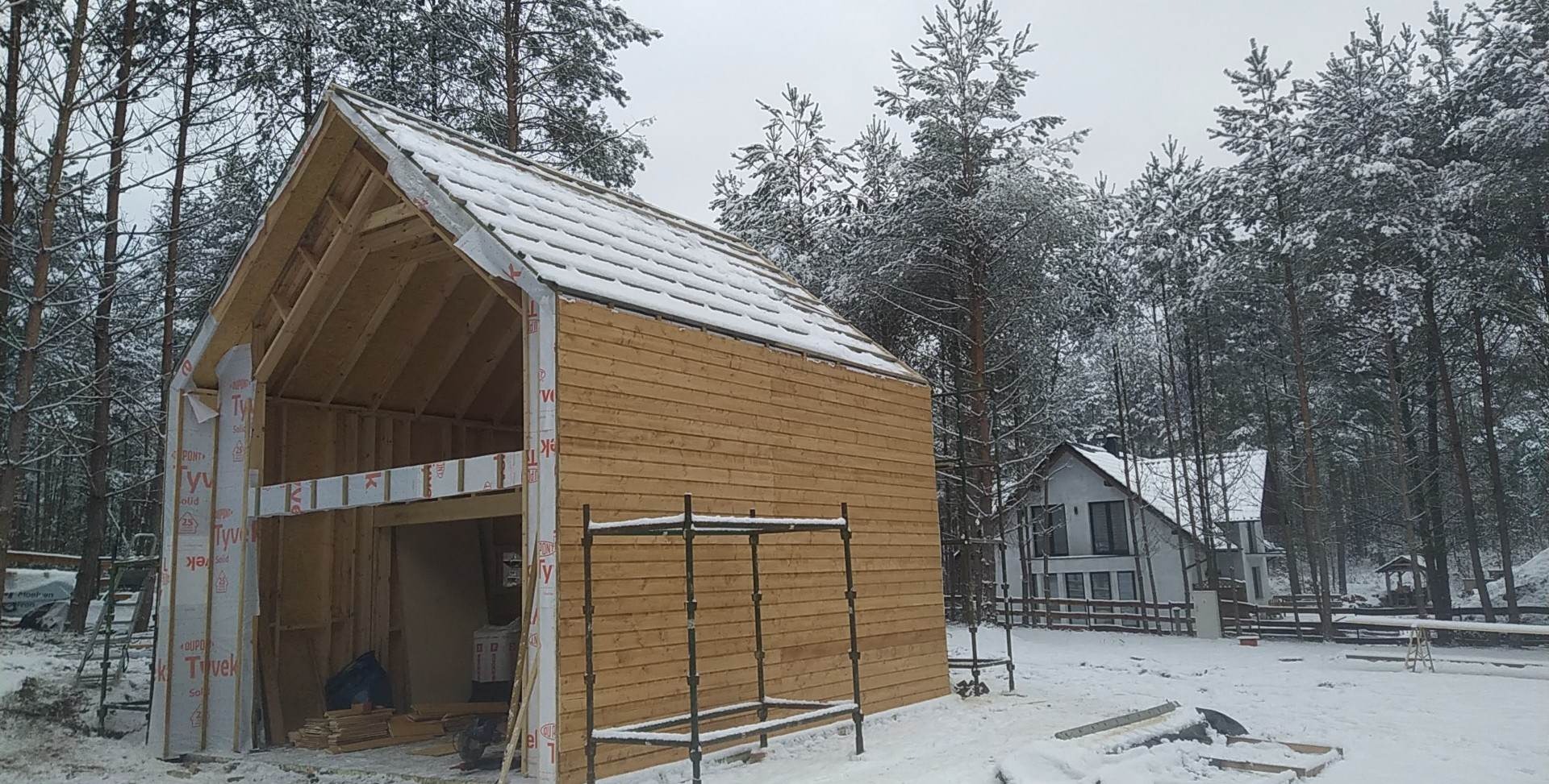 Wznoszona drewniana konstrukcja z częściowo ukończoną elewacją na tle zimowego krajobrazu. Widoczne rusztowania i materiały budowlane.