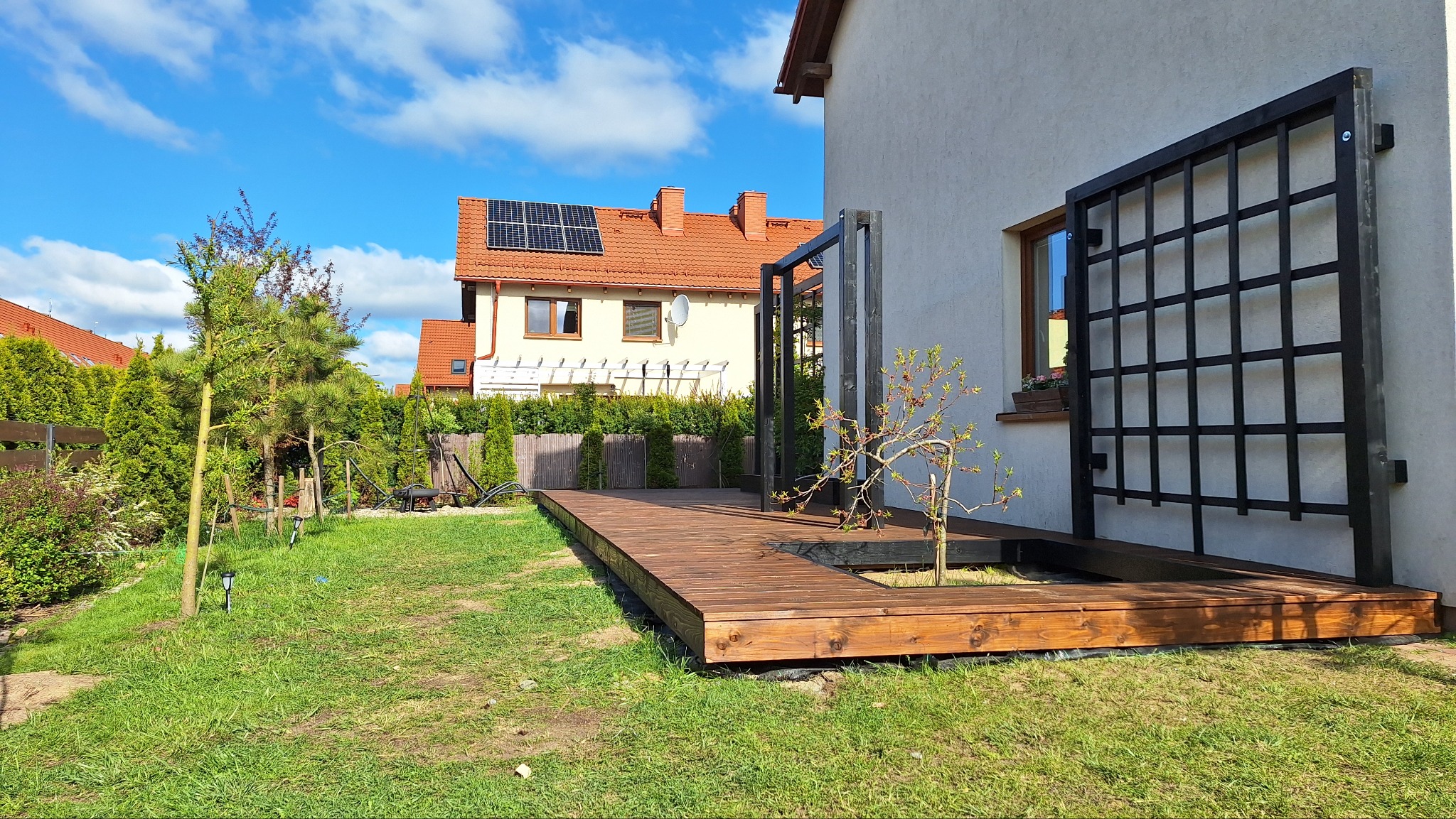 Nowoczesny taras drewniany z czarną, metalową pergolą przy ścianie domu. Widoczne drzewko wbudowane w taras i zadbany trawnik. Słoneczny dzień.