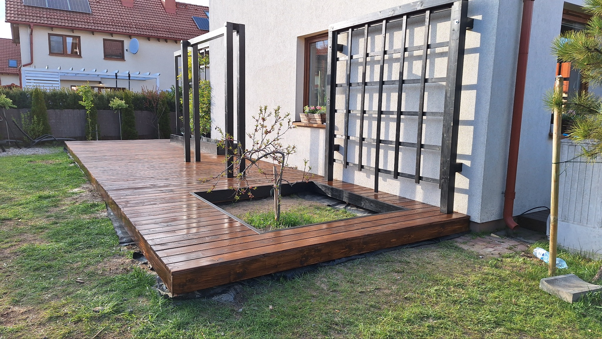 Nowoczesny taras drewniany z pergolą w formie kraty przy białym domu. Wbudowana donica z drzewkiem. Widok na ogród i sąsiedni budynek z panelami słonecznymi.