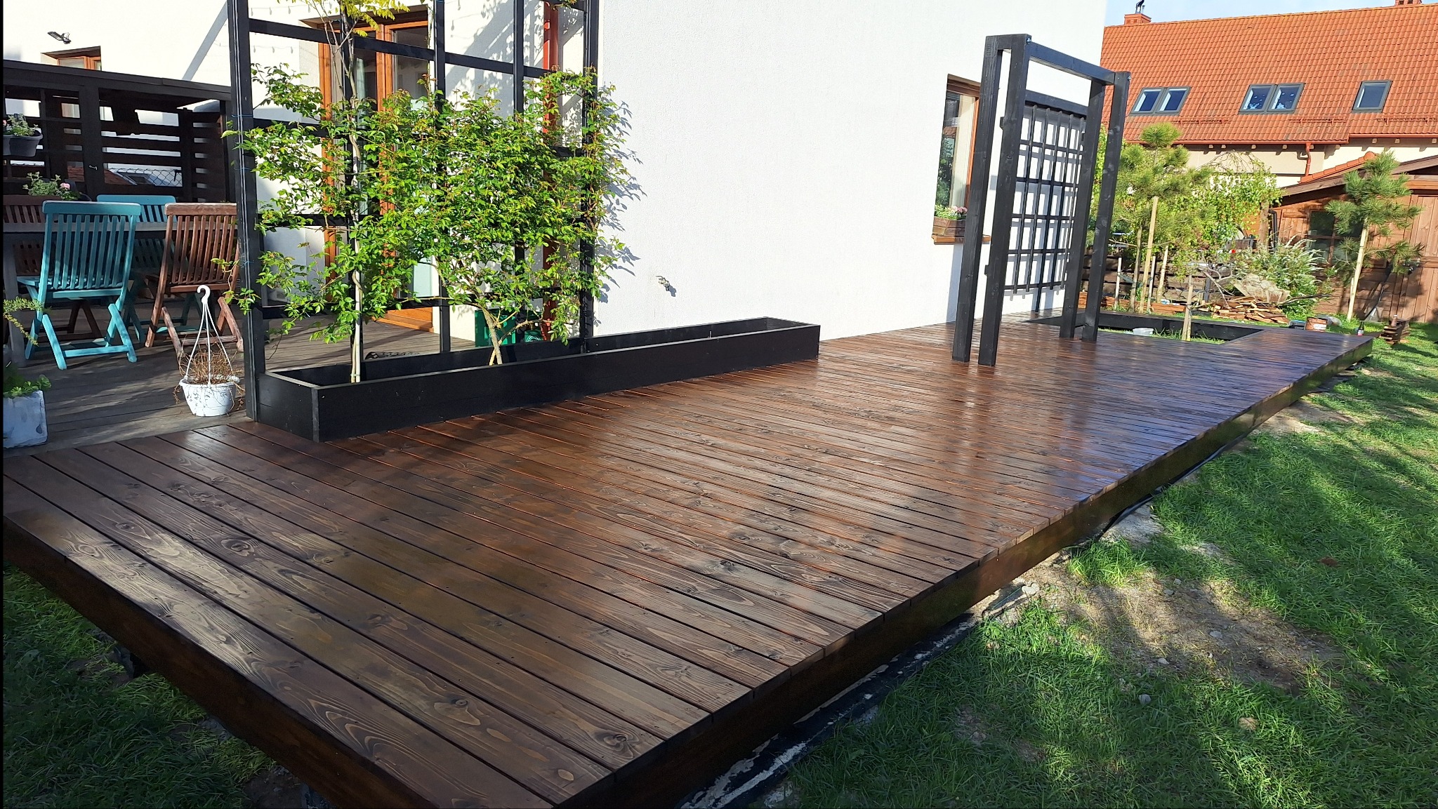 Ciemnobrązowy taras drewniany z czarną pergolą i donicą, przylegający do białego domu. Widoczne meble ogrodowe i zieleń trawnika.