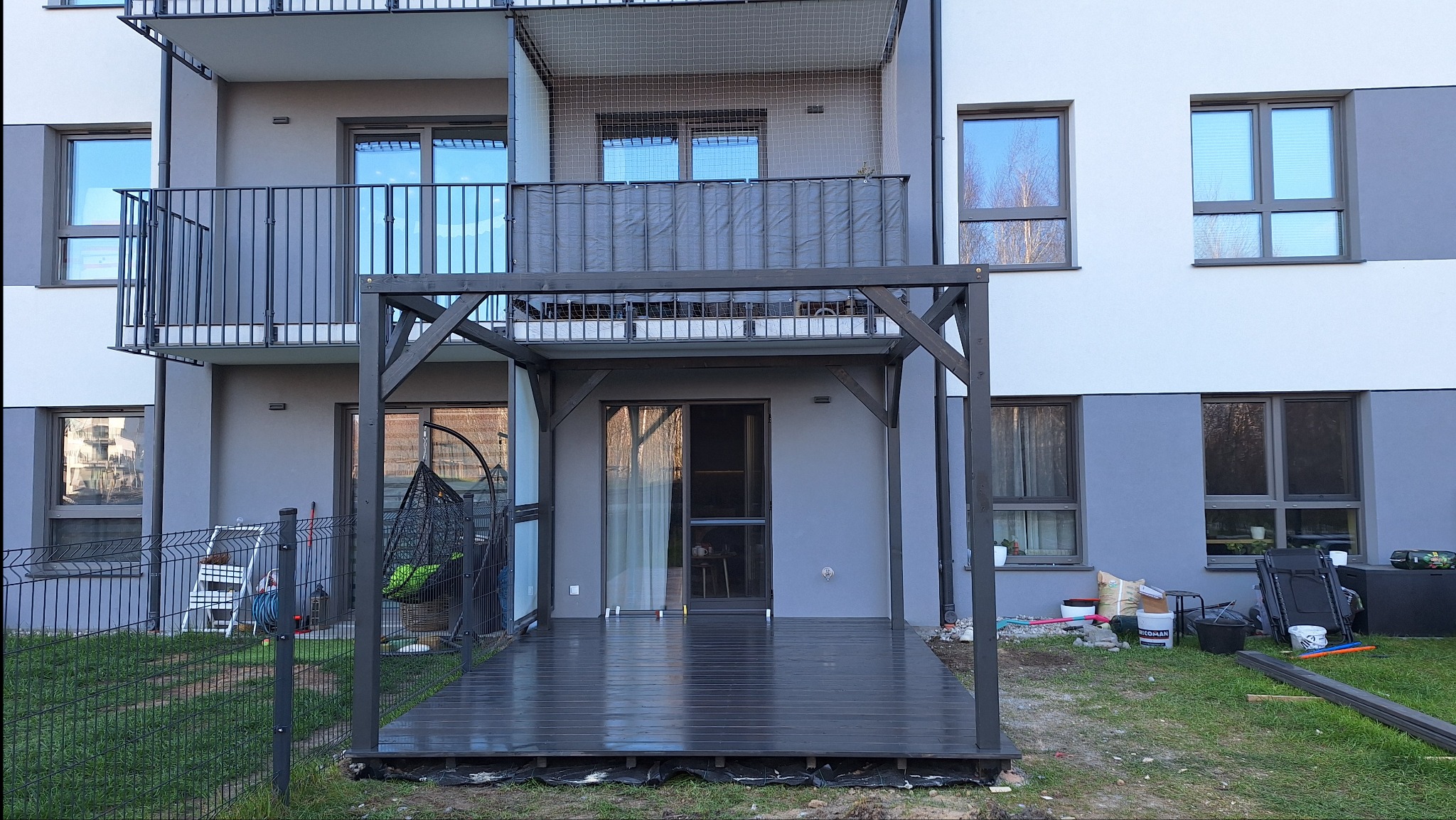 Ciemna, drewniana pergola przy balkonie w bloku, z widocznym tarasem. W tle narzędzia i materiały budowlane, sugerujące etap wykończeniowy. Nowoczesny design.