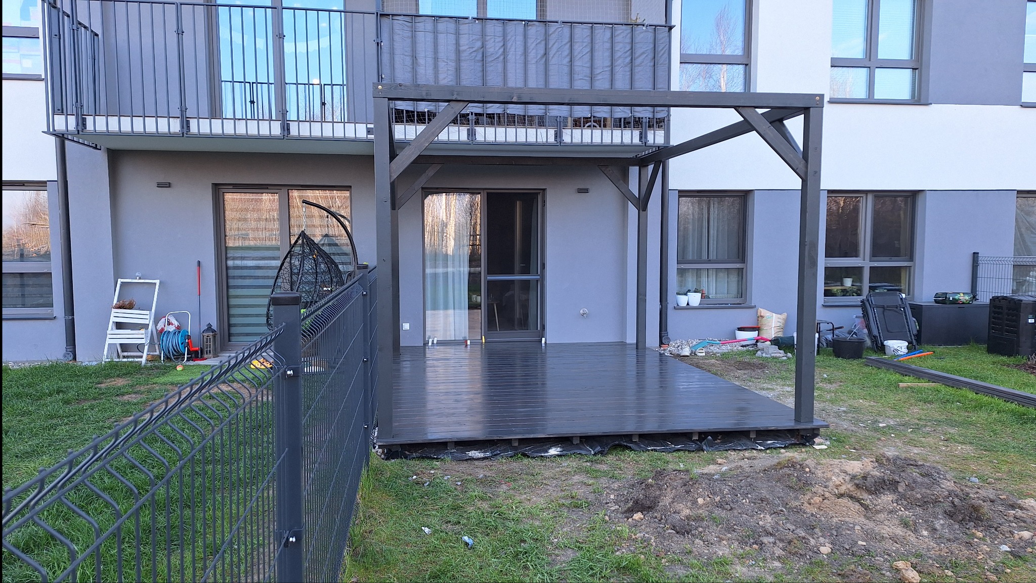 Nowoczesny taras z drewnianą pergolą w szarym kolorze, przylegający do budynku z balkonem. Widoczne ogrodzenie i elementy wykończeniowe.