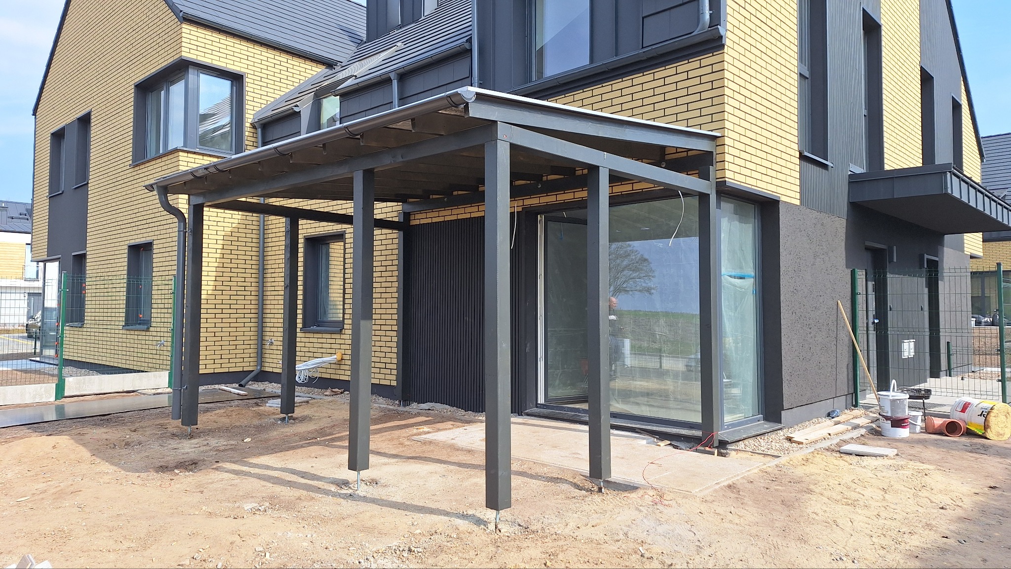 Nowoczesna, grafitowa pergola przy domu z żółtej cegły. Widoczne słupy pergoli przed montażem podłoża. Elementy wykończeniowe i narzędzia obok.