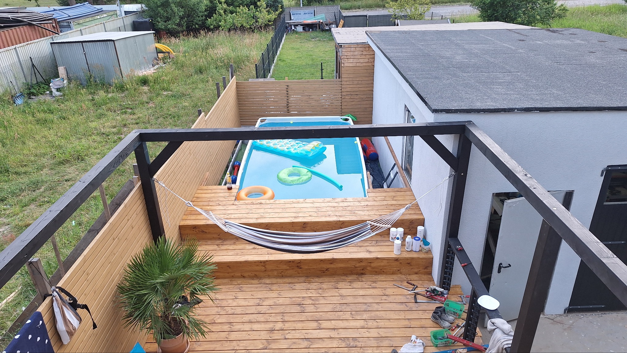 Drewniany taras z pergolą w kolorze czarnym, hamakiem i basenem. Widok z góry ukazuje detale desek, roślinę w donicy i akcesoria basenowe.