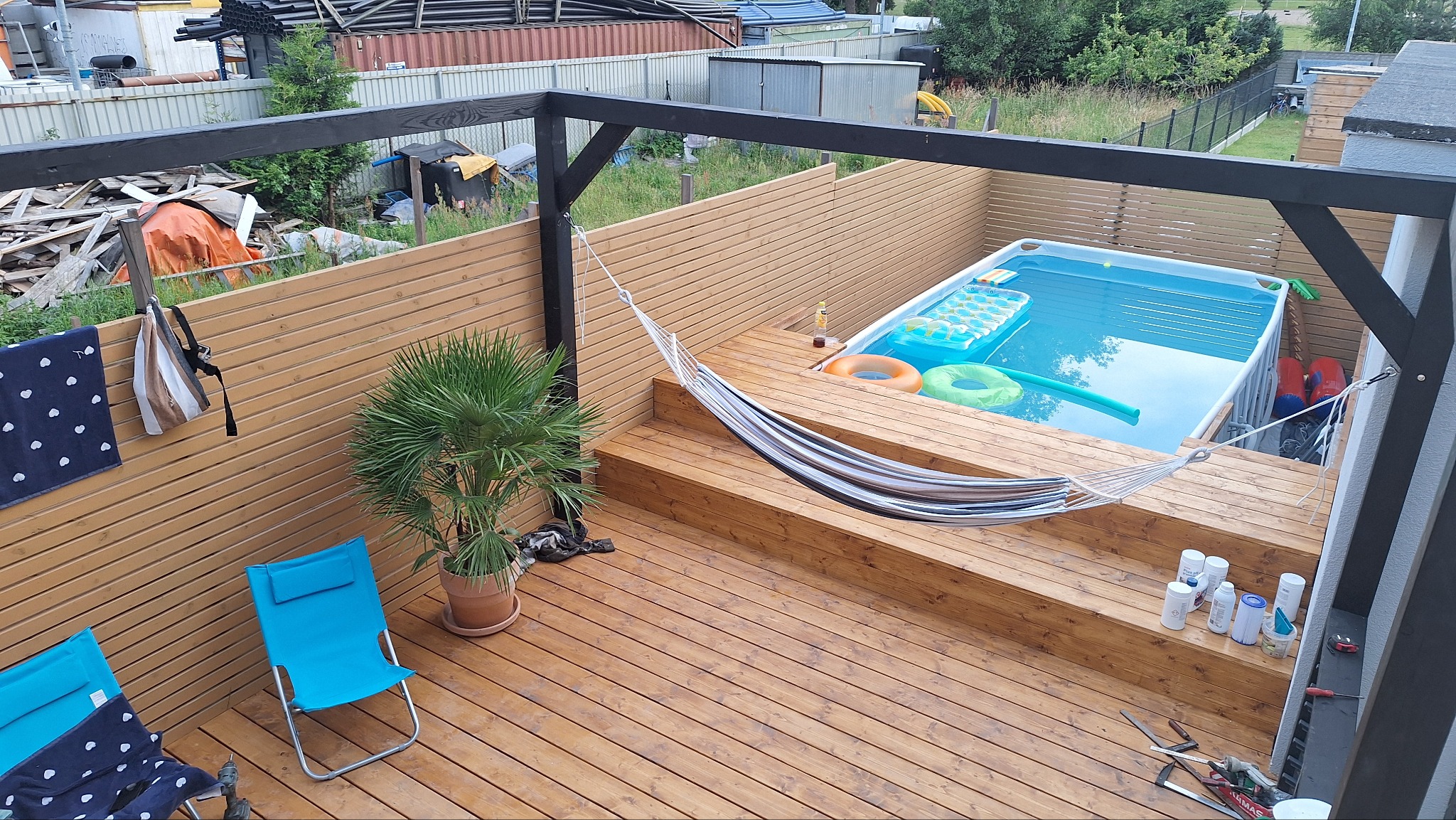 Drewniany taras z basenem i hamakiem, otoczony płotem z desek. Widoczne krzesła plażowe, roślina w doniczce i akcesoria basenowe. Czarna konstrukcja pergoli nad tarasem.