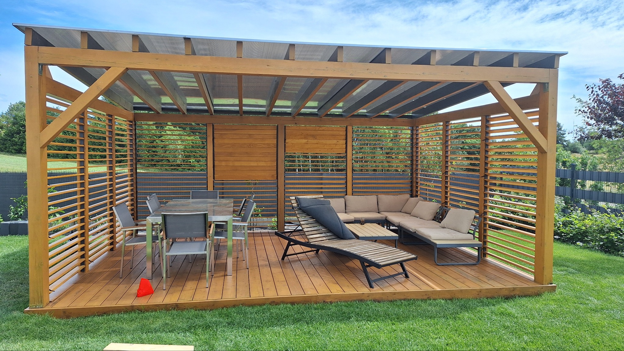 Drewniana pergola z zadaszeniem i ażurowymi ścianami, wyposażona w meble ogrodowe: stół z krzesłami, leżak i narożnik. Całość na drewnianym tarasie, otoczona zielenią.