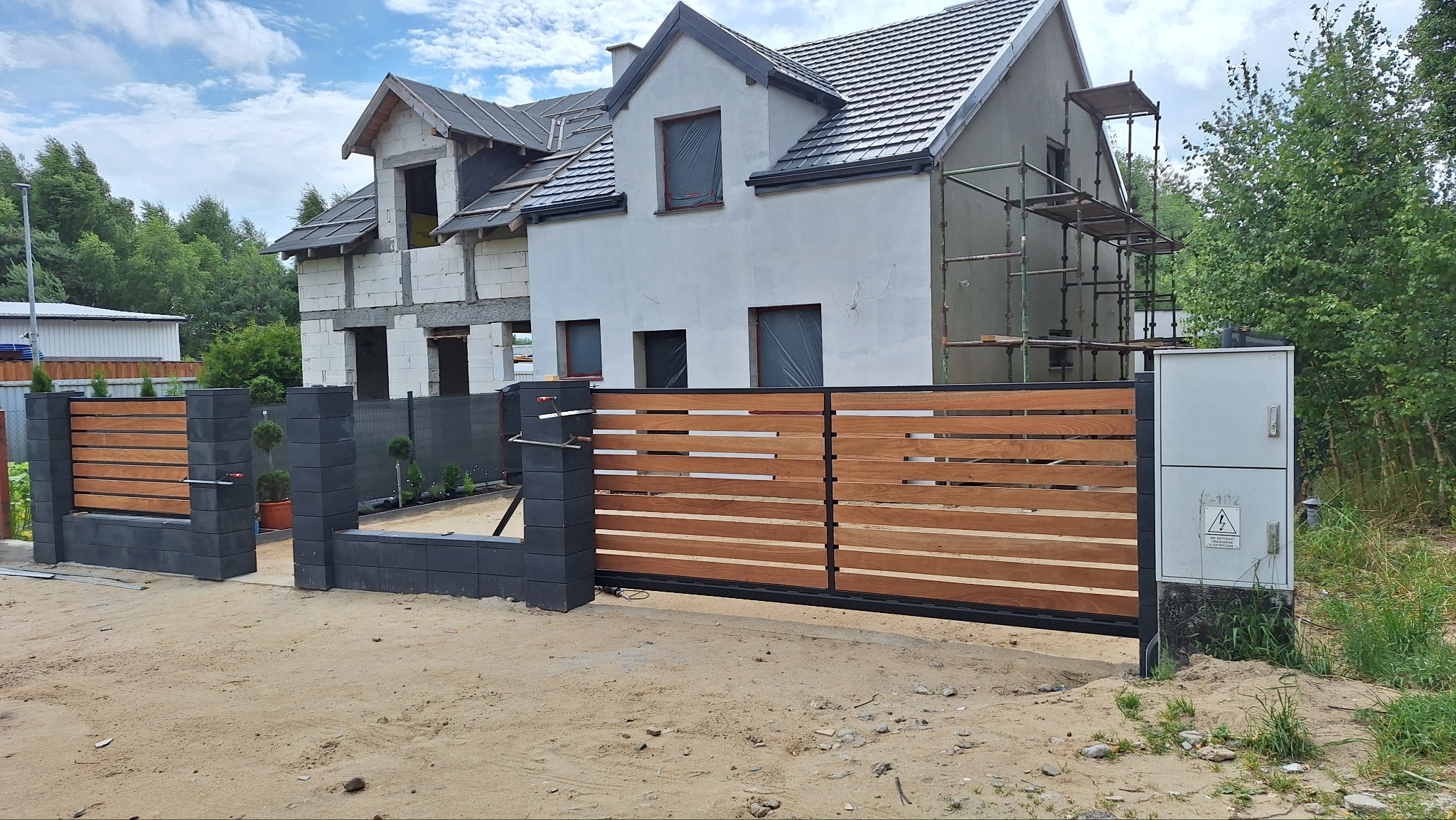 Nowoczesna brama przesuwna z drewnianymi panelami w Gdyni. Brama zamontowana na tle domu w trakcie budowy, z widocznymi rusztowaniami i elementami konstrukcyjnymi.