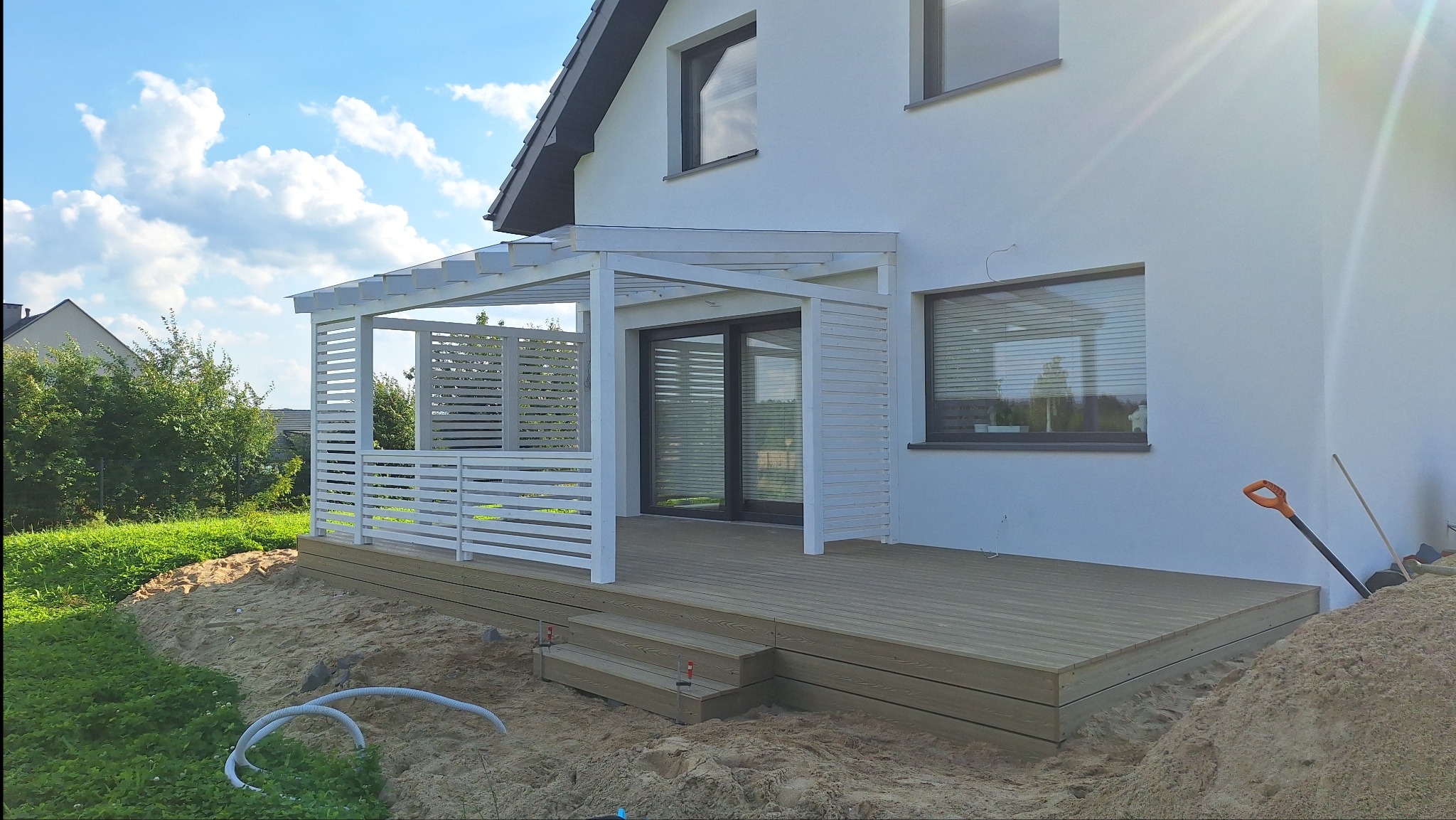 Nowoczesny taras drewniany z białą pergolą przy domu jednorodzinnym. Widoczne schody, deski tarasowe i elementy konstrukcyjne pergoli. W tle błękitne niebo i zieleń.