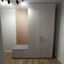 Nowoczesna szafa wnękowa w odcieniach szarości z siedziskiem i dekoracyjną ścianką z drewnianych listewek. Minimalistyczne uchwyty, oświetlenie sufitowe.