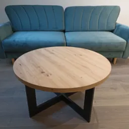 Turkusowa sofa z okrągłym stolikiem kawowym z drewnianym blatem i czarną podstawą. Minimalistyczny design, jasne wnętrze, parkiet w odcieniach szarości.