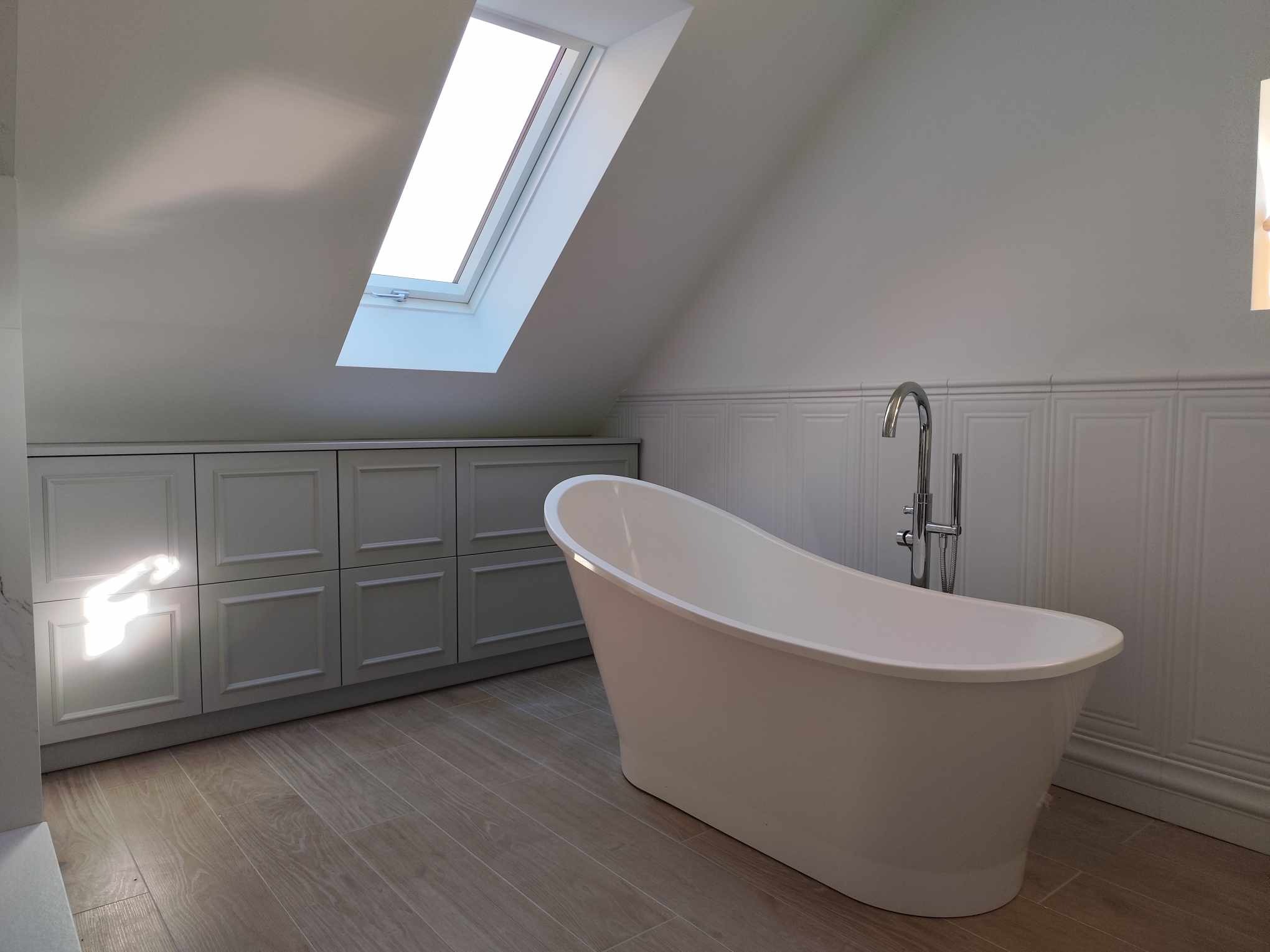 Jasna łazienka na poddaszu z wolnostojącą wanną i szafkami pod skosem, oświetlona przez okno dachowe. Minimalistyczny design i drewnopodobne płytki podłogowe.