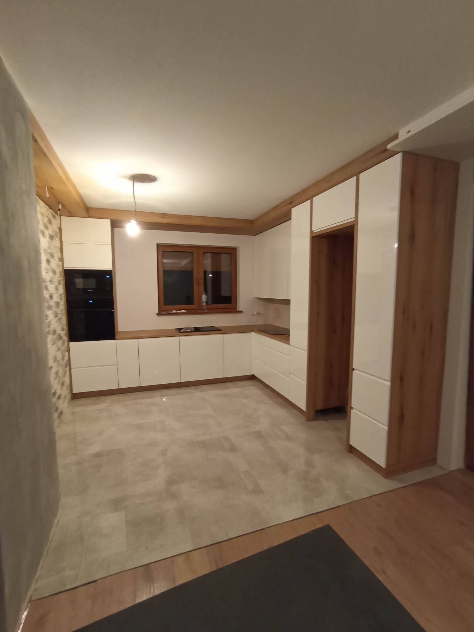 Jasna kuchnia z drewnianymi belkami stropowymi i frontami szafek w kolorze białym. Widoczne zabudowane sprzęty AGD i okno. Całość w stylu nowoczesnym.