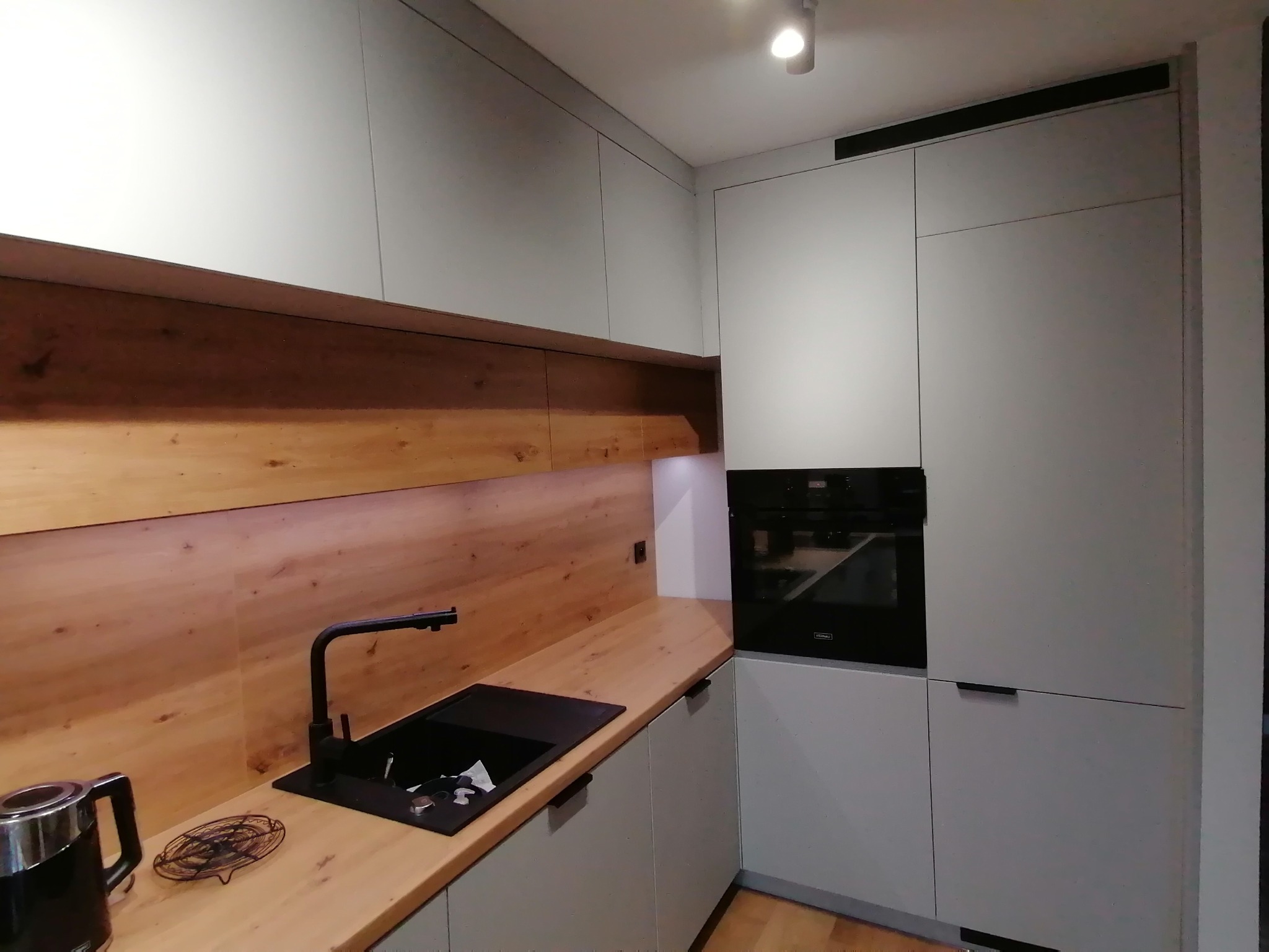 Jasnoszara kuchnia z drewnianym blatem i czarną armaturą. Uwagę przykuwa drewniana ściana nad blatem, dodająca ciepła nowoczesnemu wnętrzu.