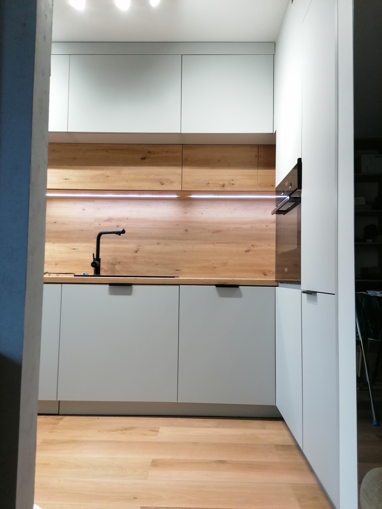 Nowoczesna kuchnia: białe szafki, drewniany panel, czarna armatura. Minimalistyczny design z oświetleniem LED. Widok zza framugi drzwi.