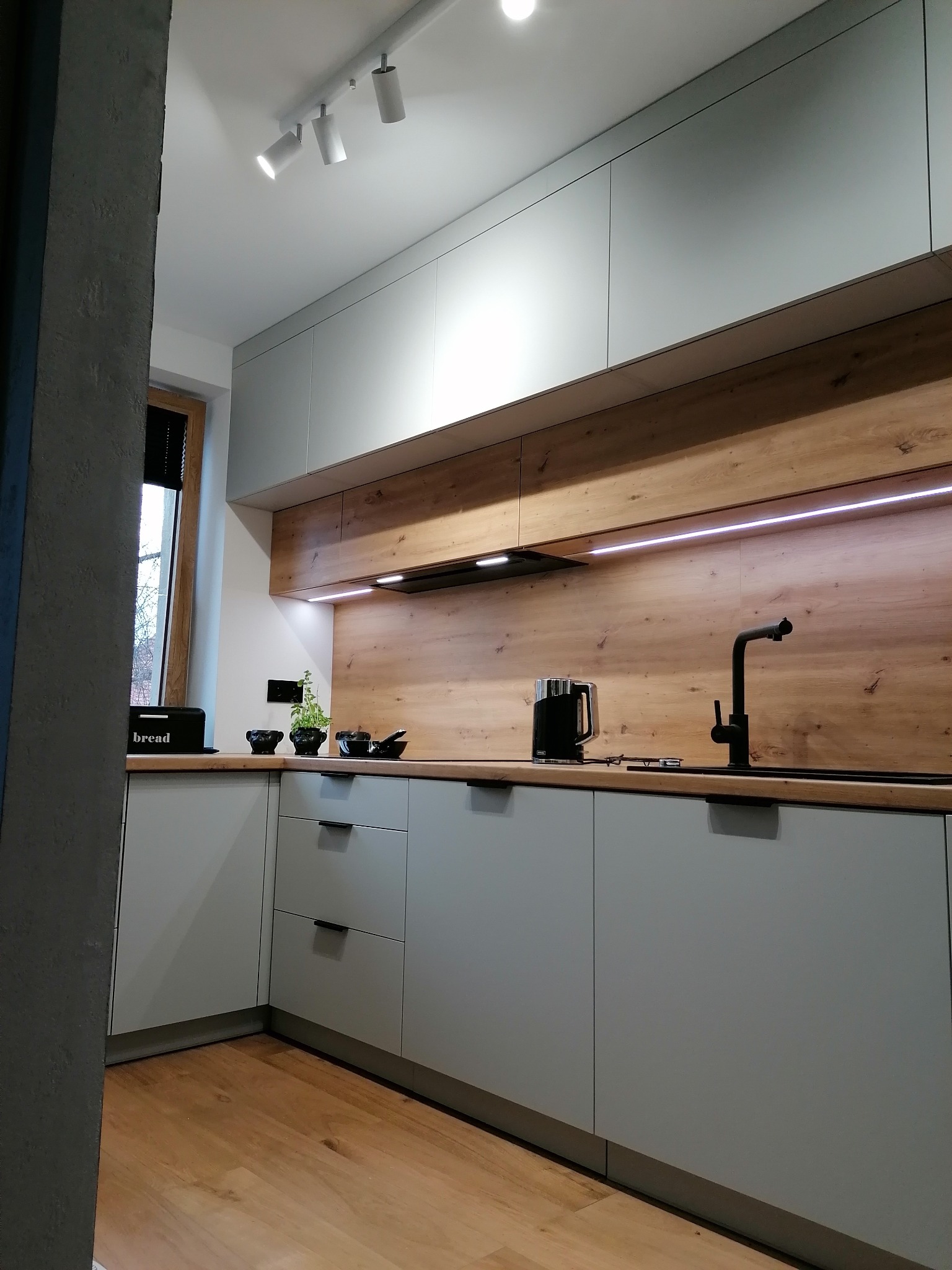 Nowoczesna kuchnia z szarymi frontami i drewnianym panelem ściennym z oświetleniem LED. Czarny kran i akcesoria kuchenne na blacie. Minimalistyczny design.