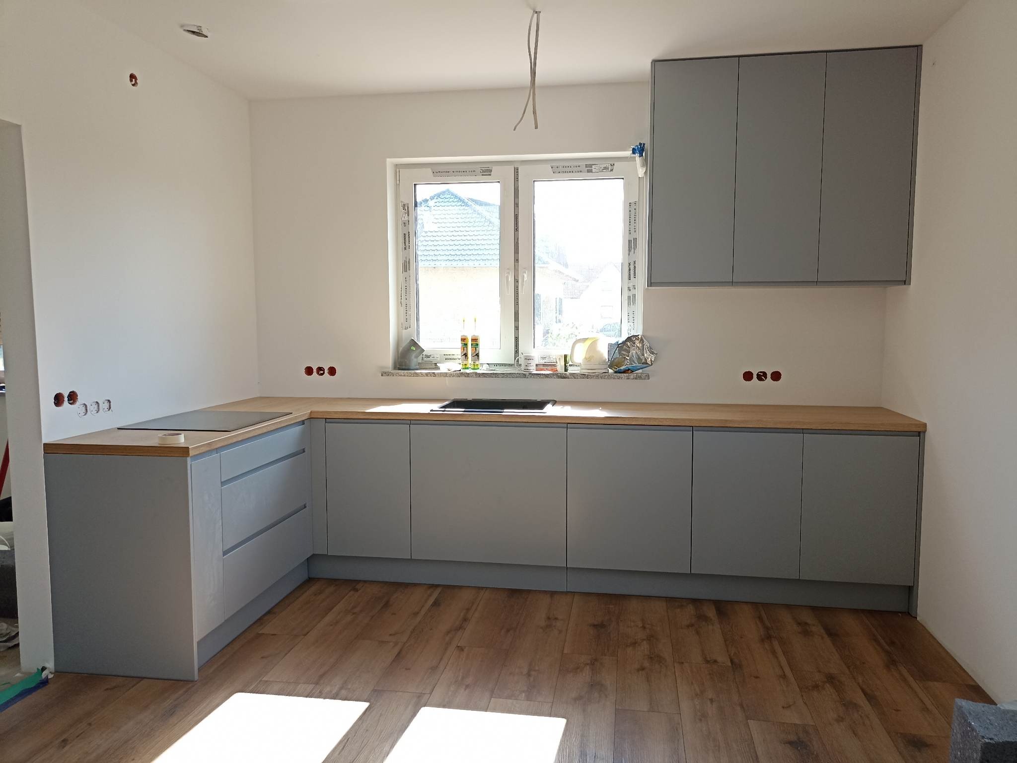 Szara kuchnia z drewnianym blatem w Wołowie. Górne szafki wiszące, dolne z szufladami. Widoczne gniazdka elektryczne i okno z widokiem na zewnątrz.