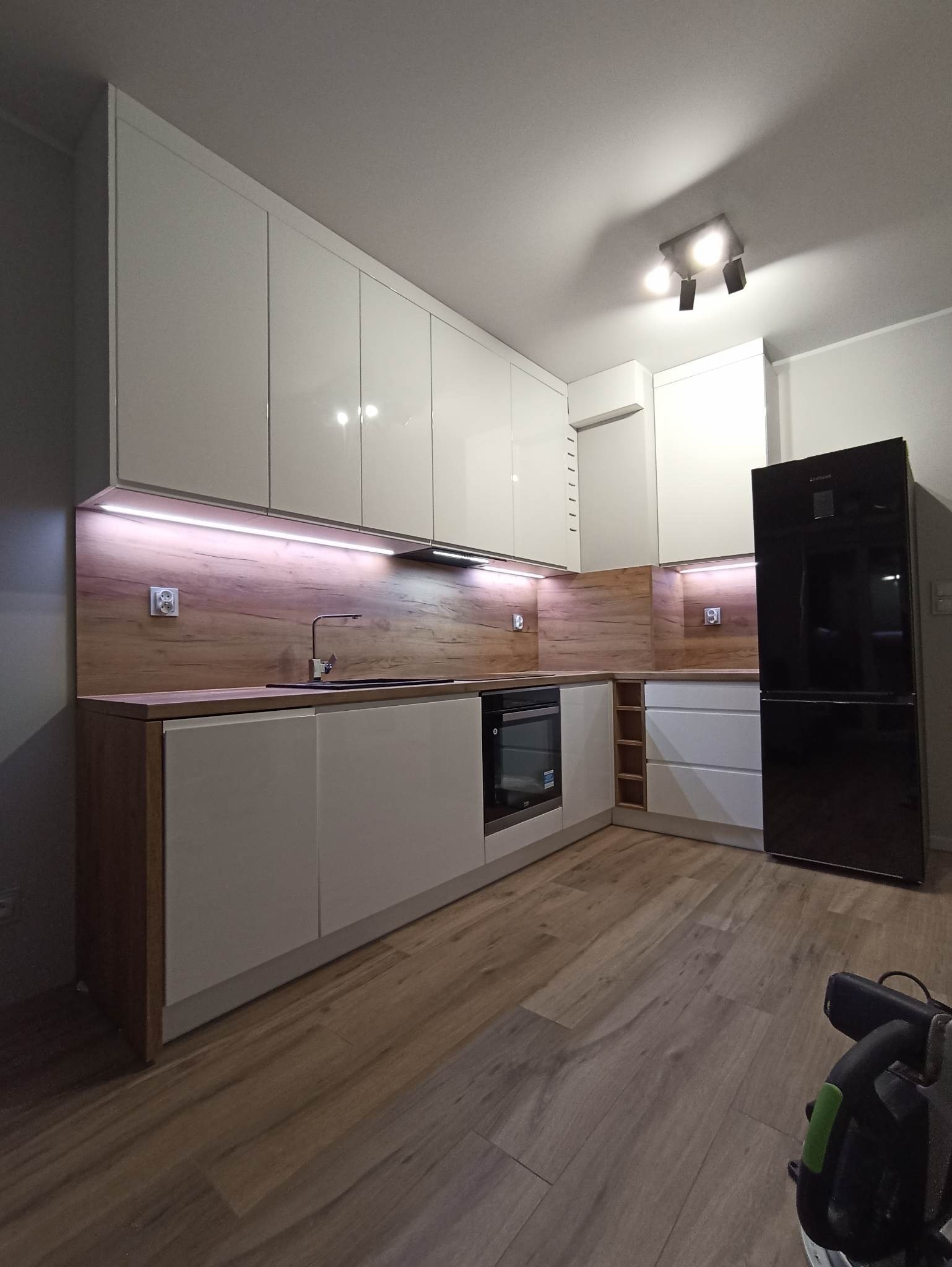 Nowoczesna kuchnia z białymi, lakierowanymi frontami, drewnianym blatem i oświetleniem LED pod szafkami wiszącymi. Czarna lodówka wolnostojąca w rogu.