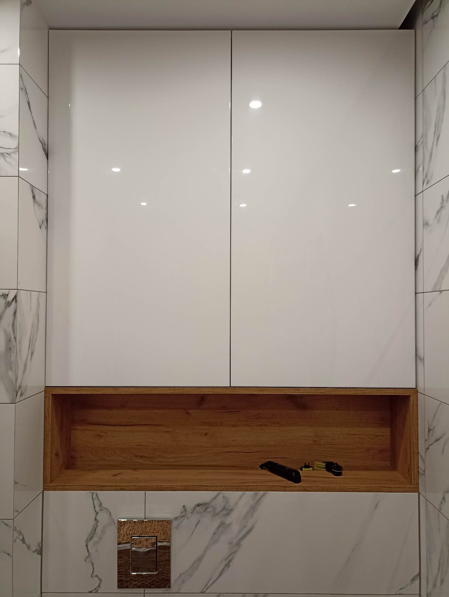 Biała szafka łazienkowa z drewnianą wnęką, w której leży trymer. Marmurowe płytki na ścianie. Nowoczesny design.