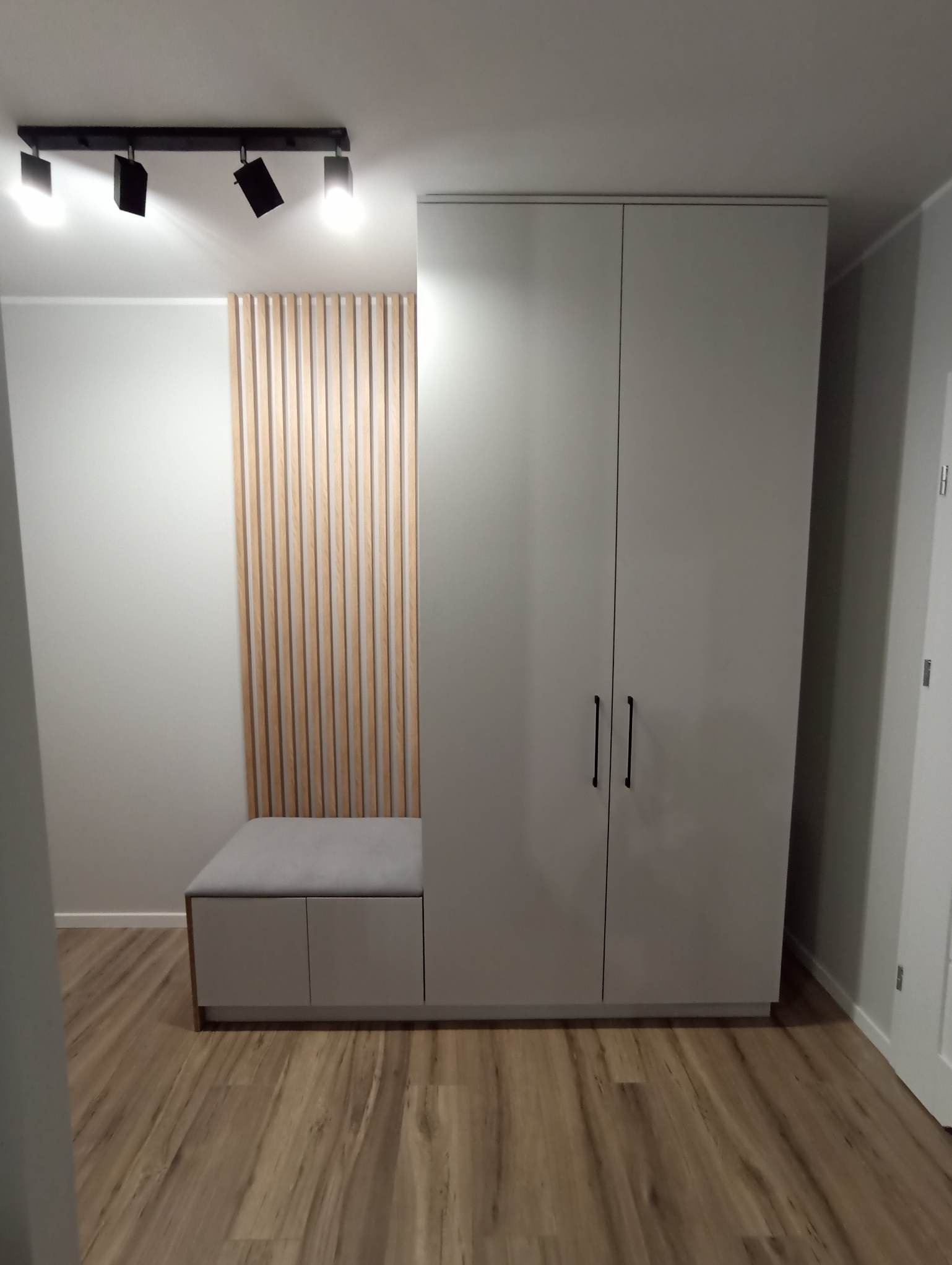 Nowoczesna szafa wnękowa w odcieniach szarości z siedziskiem i dekoracyjną ścianką z drewnianych listewek. Minimalistyczne uchwyty, oświetlenie sufitowe.
