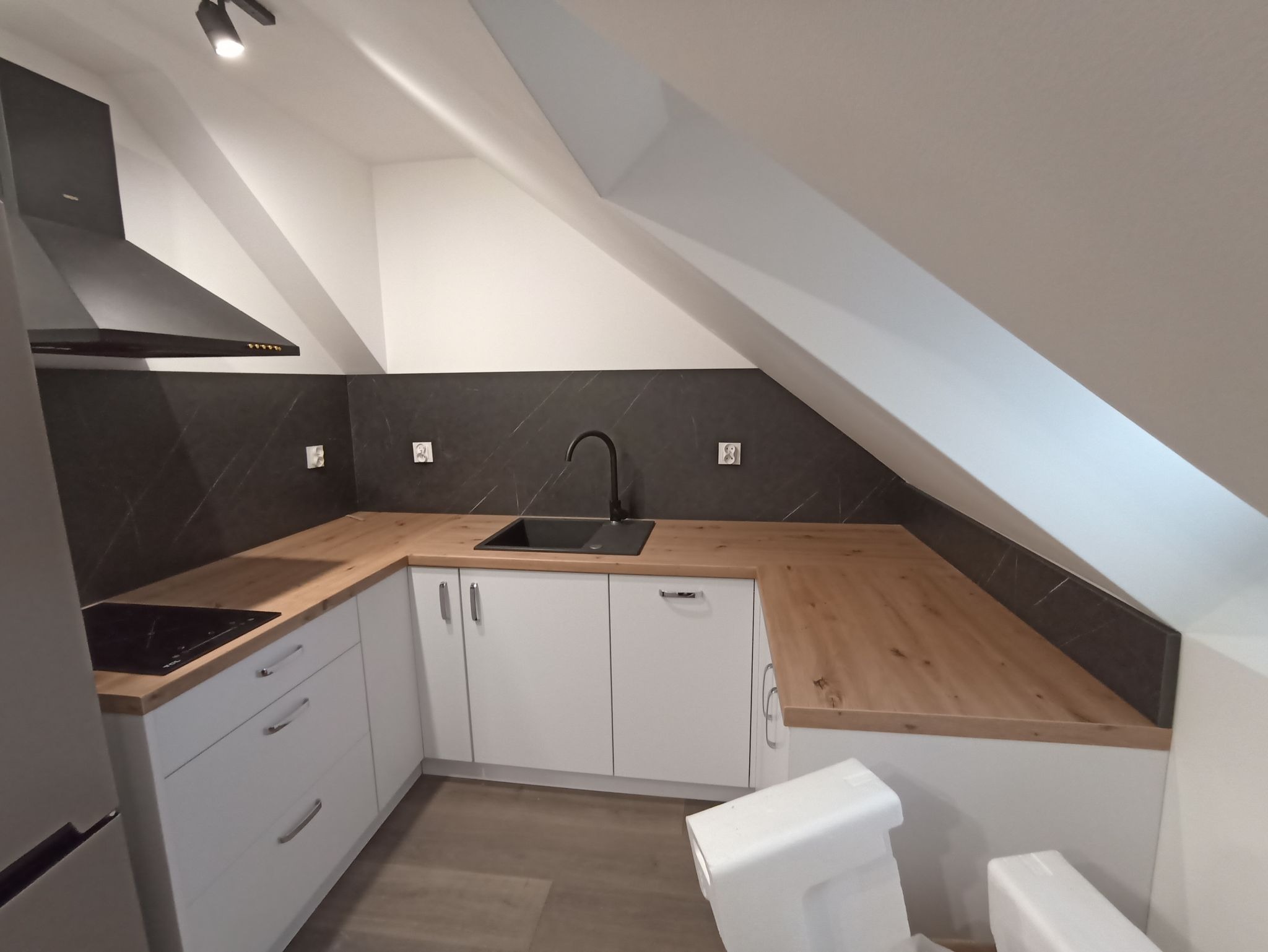 Nowoczesna kuchnia na poddaszu z białymi szafkami, drewnianym blatem i czarnym panelem ściennym. Uwagę zwraca nietypowy układ pomieszczenia.