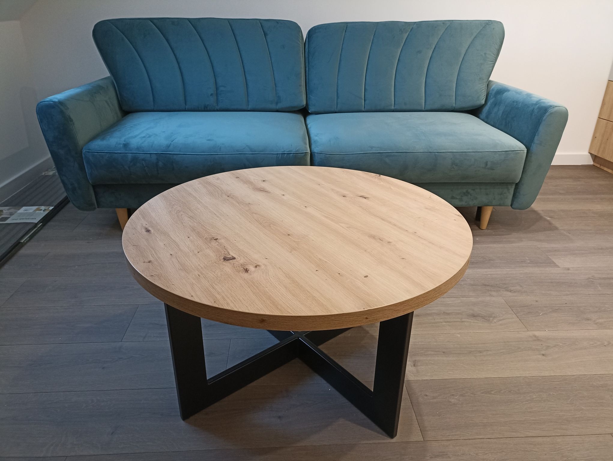 Turkusowa sofa z okrągłym stolikiem kawowym z drewnianym blatem i czarną podstawą. Minimalistyczny design, jasne wnętrze, parkiet w odcieniach szarości.