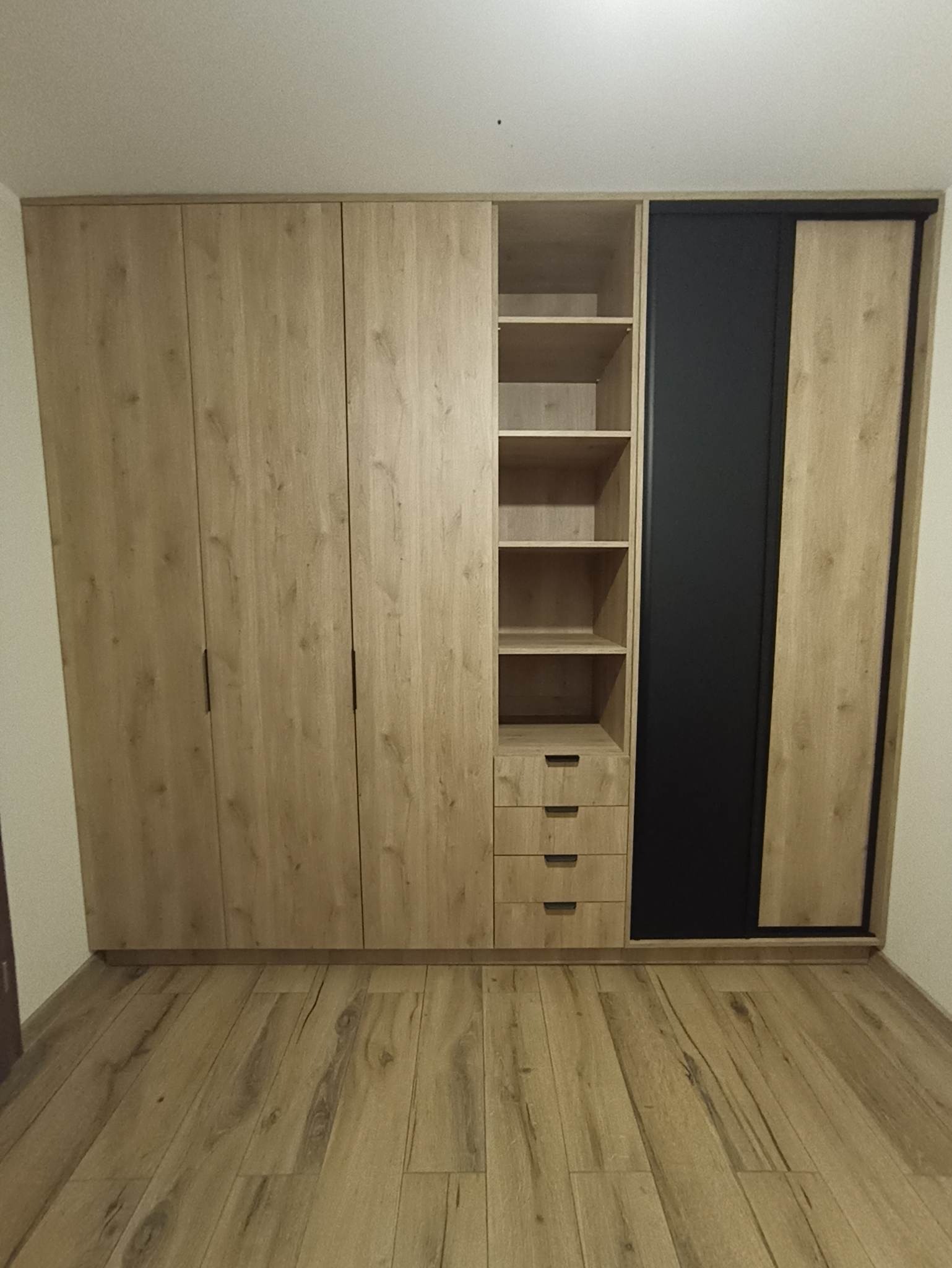Szafa na wymiar z przesuwnymi drzwiami, fronty w odcieniach drewna, czarny panel, półki i szuflady. Podłoga drewniana w jodełkę, minimalistyczny design.