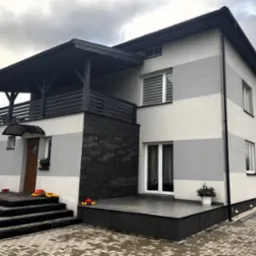 Dwukondygnacyjny dom z elewacją w pasy szarości, czarnym kamieniem dekoracyjnym i balkonem z drewnianą balustradą, ozdobiony dyniami i roślinami w donicach na schodach i tarasie.