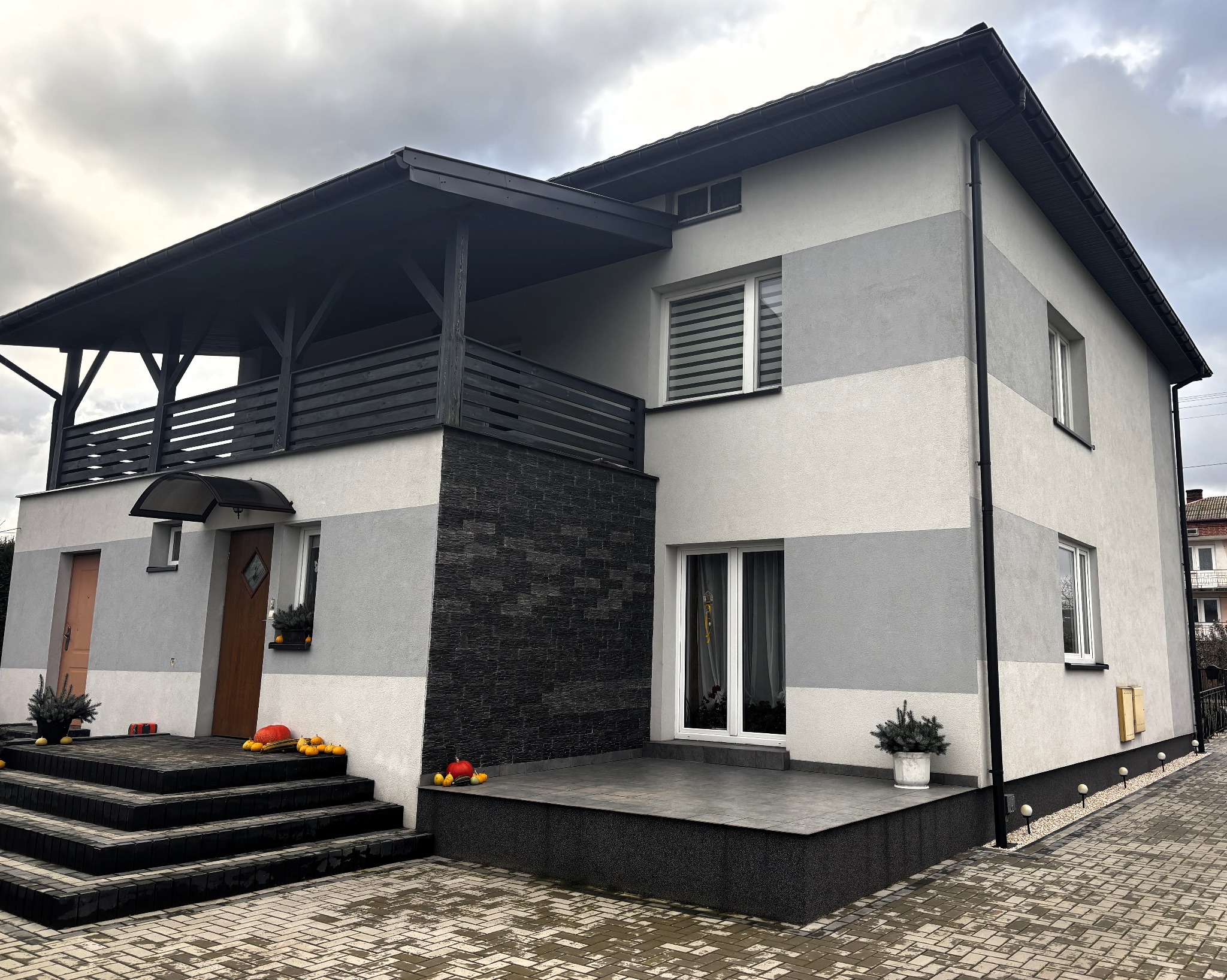 Dwukondygnacyjny dom z elewacją w pasy szarości, czarnym kamieniem dekoracyjnym i balkonem z drewnianą balustradą, ozdobiony dyniami i roślinami w donicach na schodach i tarasie.
