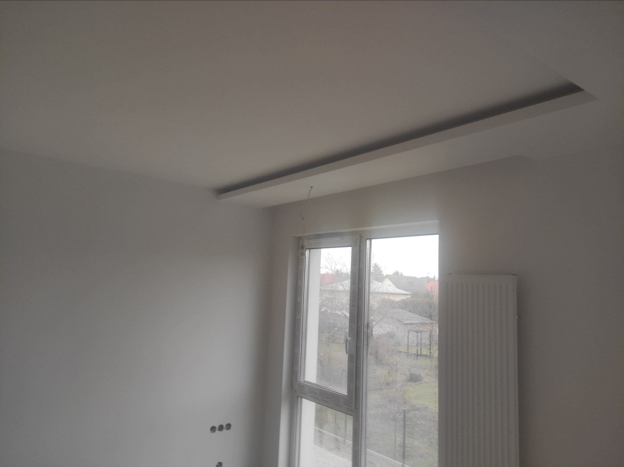 Wnętrze pokoju w stanie deweloperskim z zamontowanym oknem PCV, grzejnikiem panelowym i podwieszonym sufitem z listwą oświetleniową. Widoczne puszki elektryczne na ścianie.