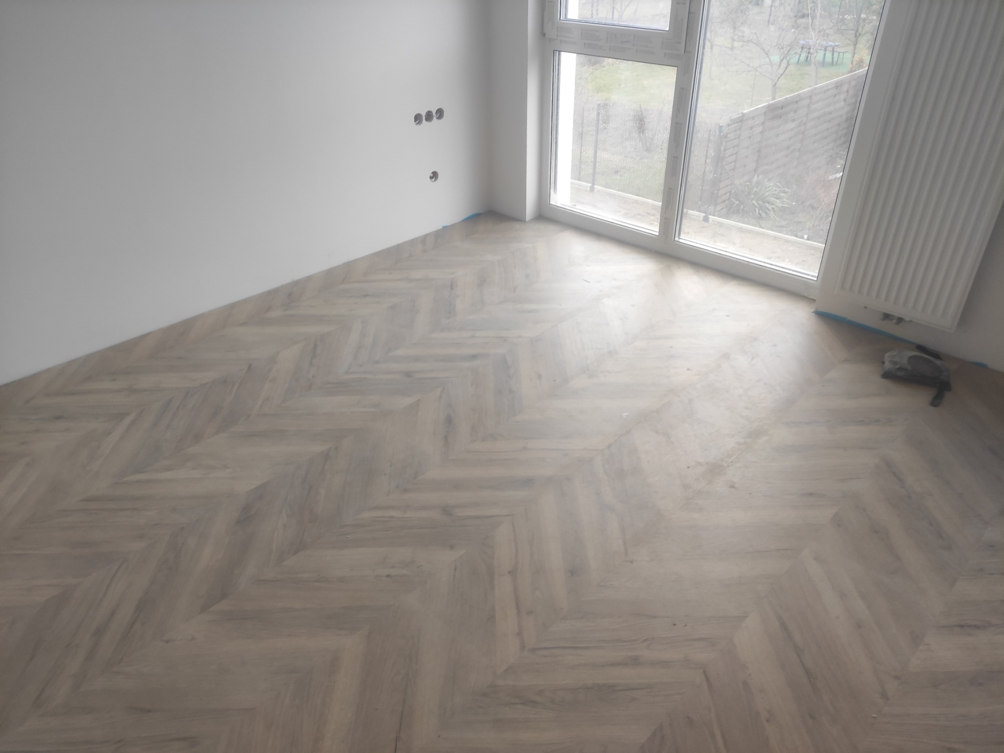 Ułożony parkiet w jodełkę w jasnym pomieszczeniu z białymi ścianami i dużym oknem, widoczne gniazdka elektryczne na ścianie, częściowo zakryty grzejnik.