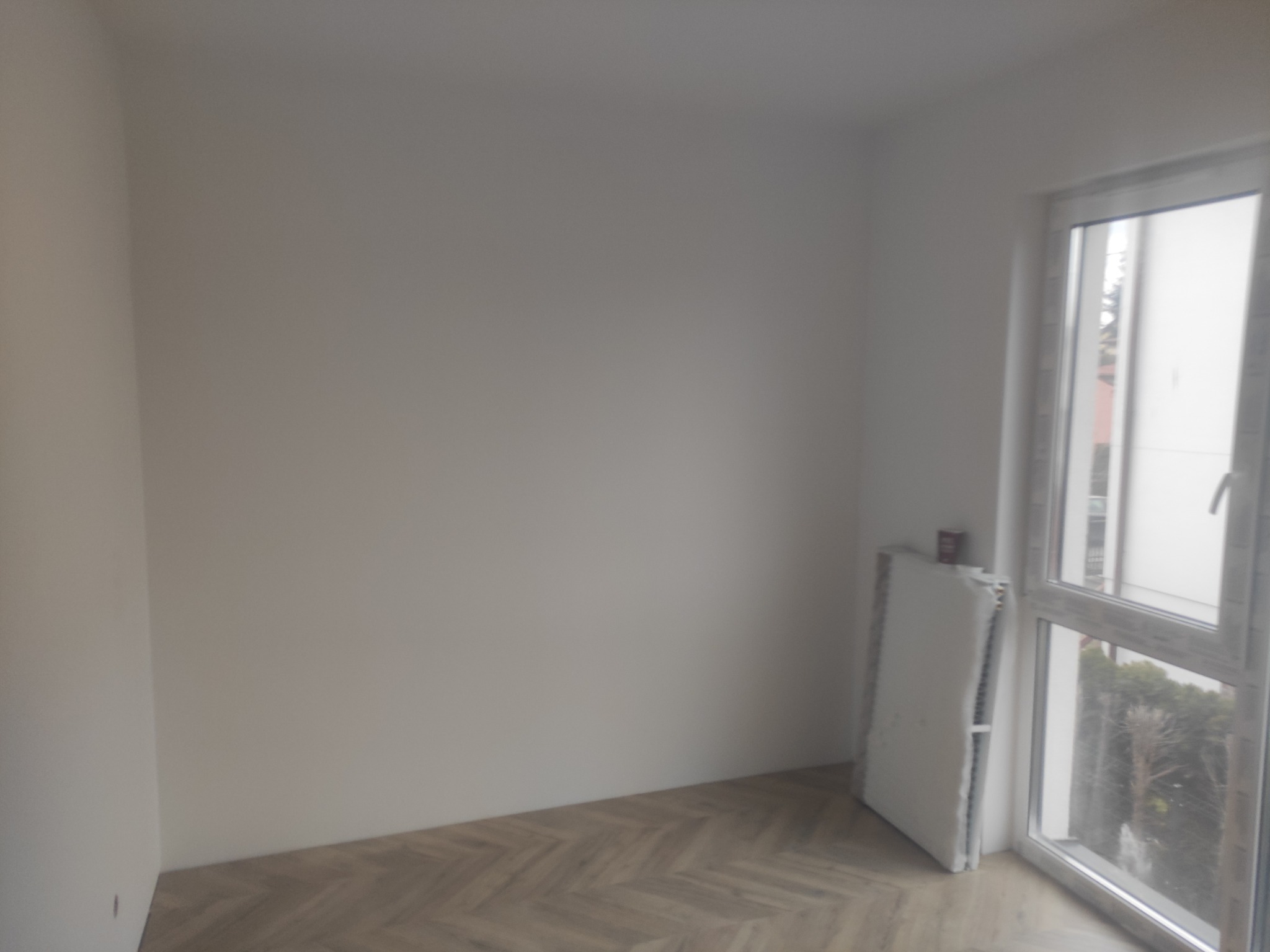 Pusty pokój z parkietem ułożonym w jodełkę, świeżo pomalowane ściany na biało, okno PCV i złożony grzejnik pod oknem