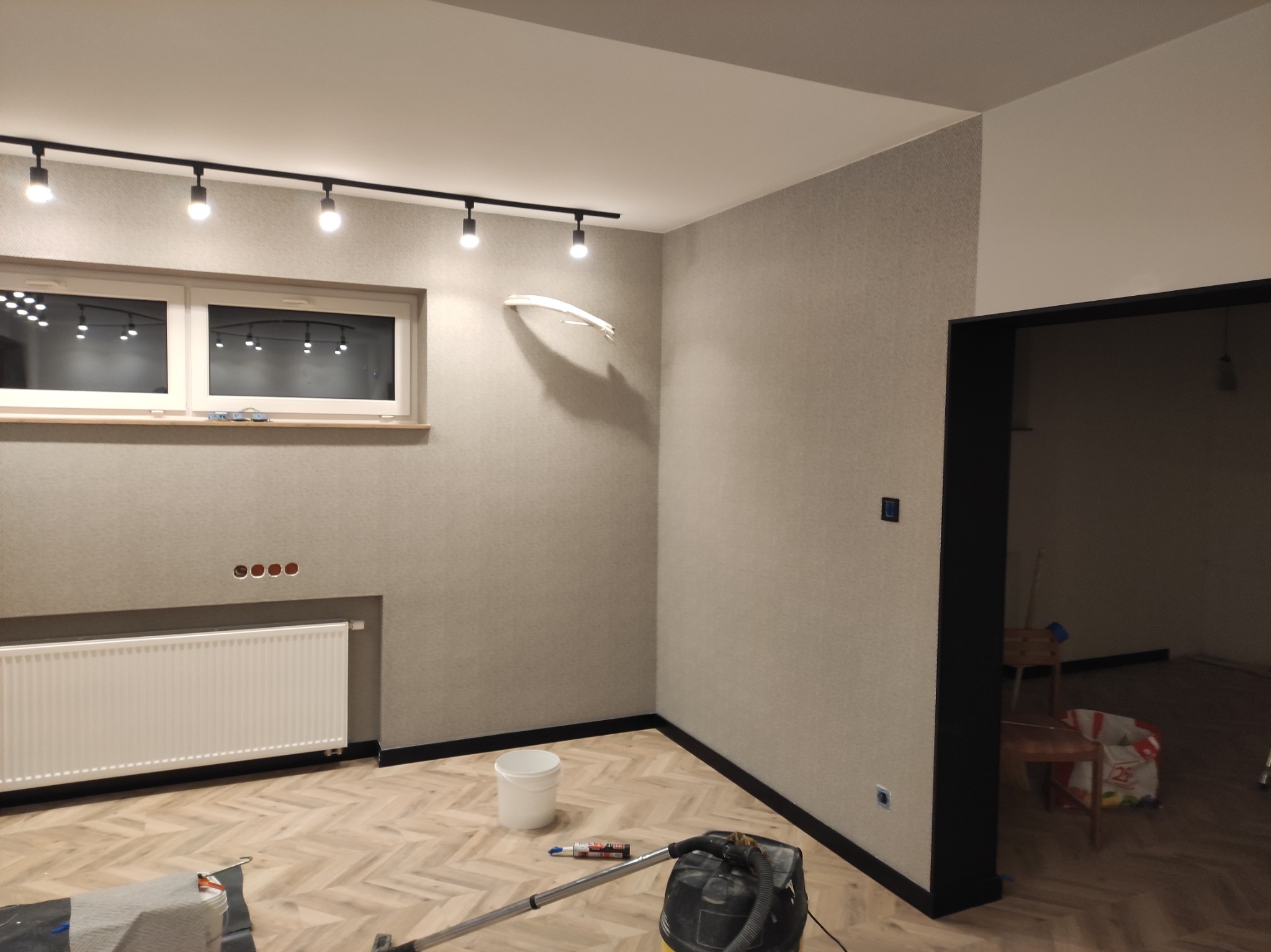 Pokój po odświeżeniu, ściany pokryte tapetą w odcieniach szarości, biały grzejnik pod oknem, na podłodze parkiet ułożony w jodełkę, widoczny biały kubeł i odkurzacz przemysłowy.