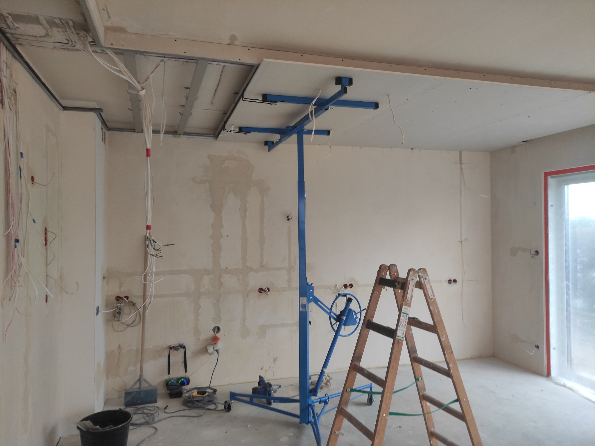 Wnętrze w trakcie prac: podwieszany sufit z płyt kartonowo-gipsowych, podnośnik do płyt w kolorze niebieskim i drewniana drabina. Widoczne wystające przewody elektryczne i przygotowane puszki...