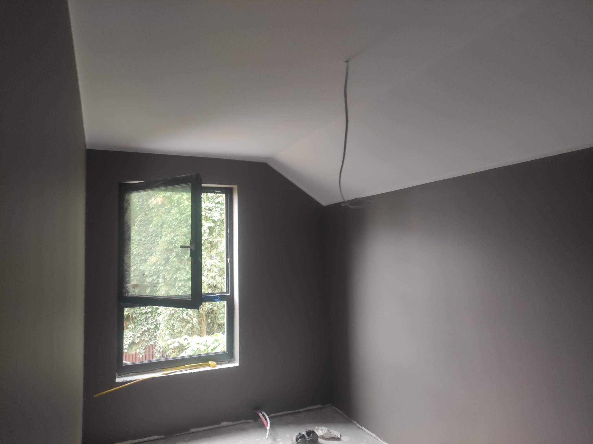 Pokój po malowaniu ścian na szaro, z otwartym oknem, przez które widać zieleń, i wystającym z sufitu przewodem elektrycznym.