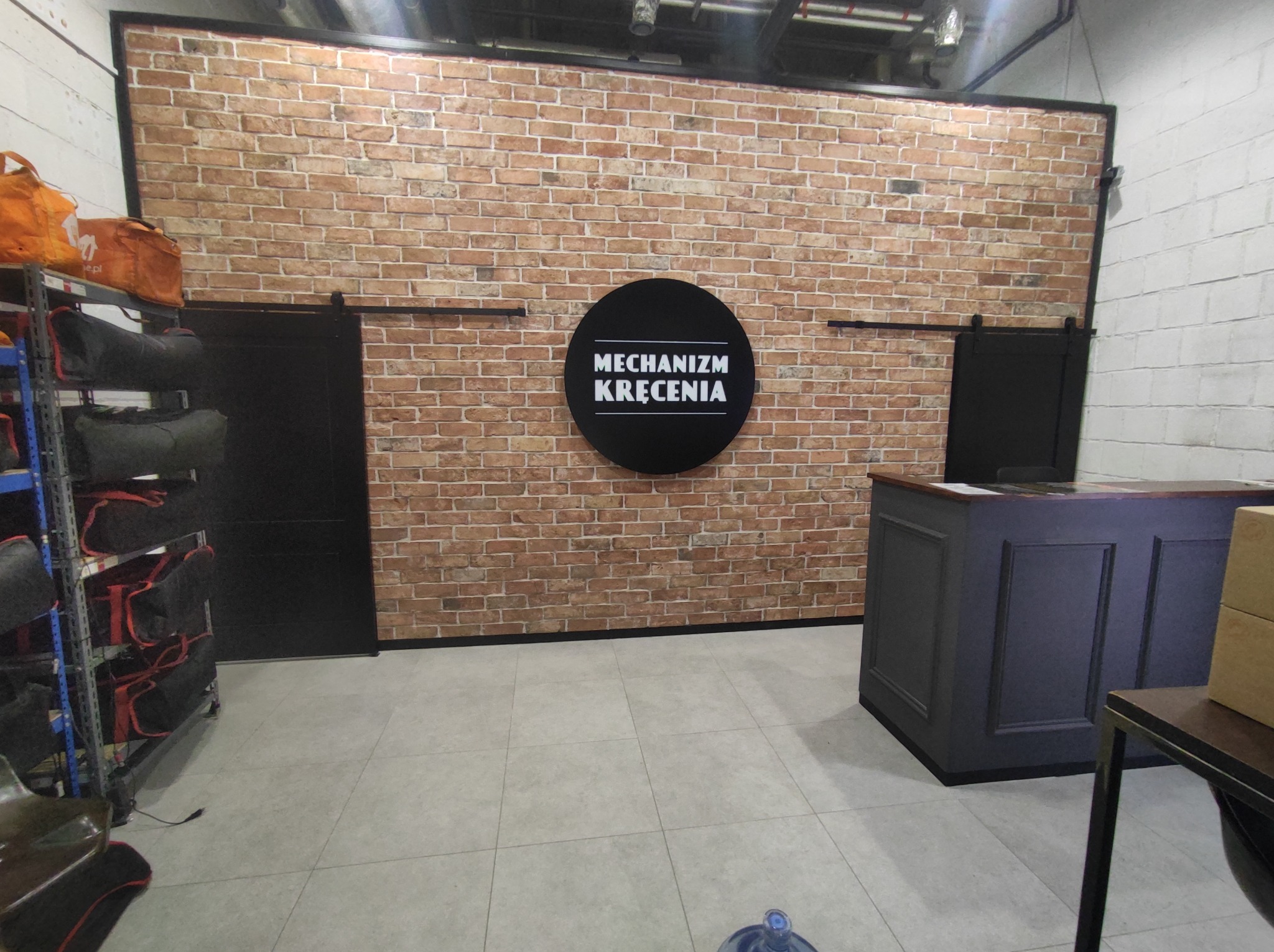 Biuro z ceglaną ścianą, czarnymi przesuwnymi drzwiami i recepcją w kolorze ciemnego grafitu, z logo 'Mechanizm Kręcenia' na ścianie.