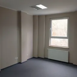 Puste biuro pomalowane na beżowo z szarym dywanem, grzejnikiem pod oknem i lampami sufitowymi.