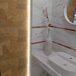 GRUPA FLEXIBLE SPÓŁKA Z OGRANICZONĄ ODPOWIEDZIALNOŚCIĄ - Nowoczesna łazienka z umywalką nablatową, drewnopodobną ścianą z podświetleniem LED i marmurowymi płytkami z dekoracyjnymi listwami, uzupełniona wazonem z bawełną.