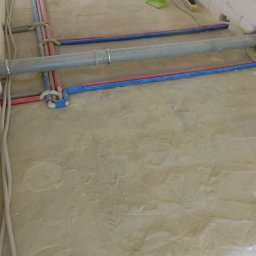 Instalacja wodno-kanalizacyjna i elektryczna w bruzdach pod wylewką. Widoczne rury PEX w kolorze niebieskim i czerwonym oraz peszle z kablami. Ściany z betonu komórkowego.
