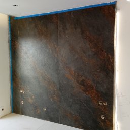 Ściana wyłożona ciemnymi płytami imitującymi kamień z widocznymi otworami na gniazdka elektryczne, zabezpieczona niebieską taśmą malarską przy suficie i ścianach.