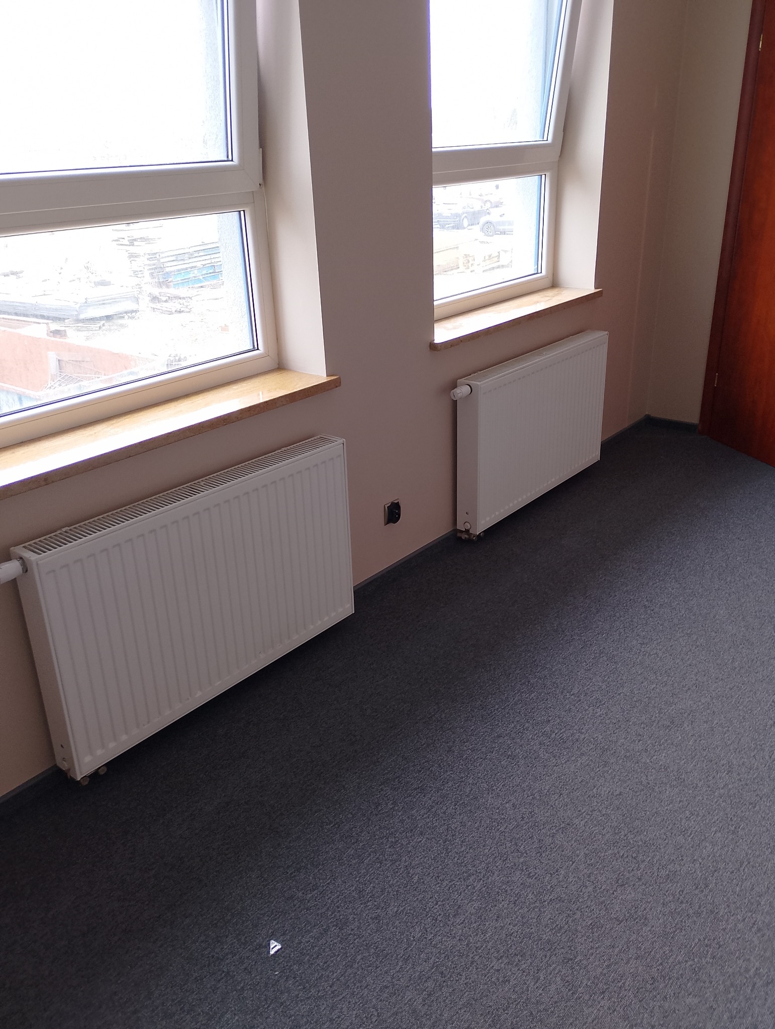 Pusta przestrzeń biurowa z dwoma białymi grzejnikami pod oknami, szara wykładzina dywanowa, beżowe ściany i częściowo widoczne drewniane drzwi.