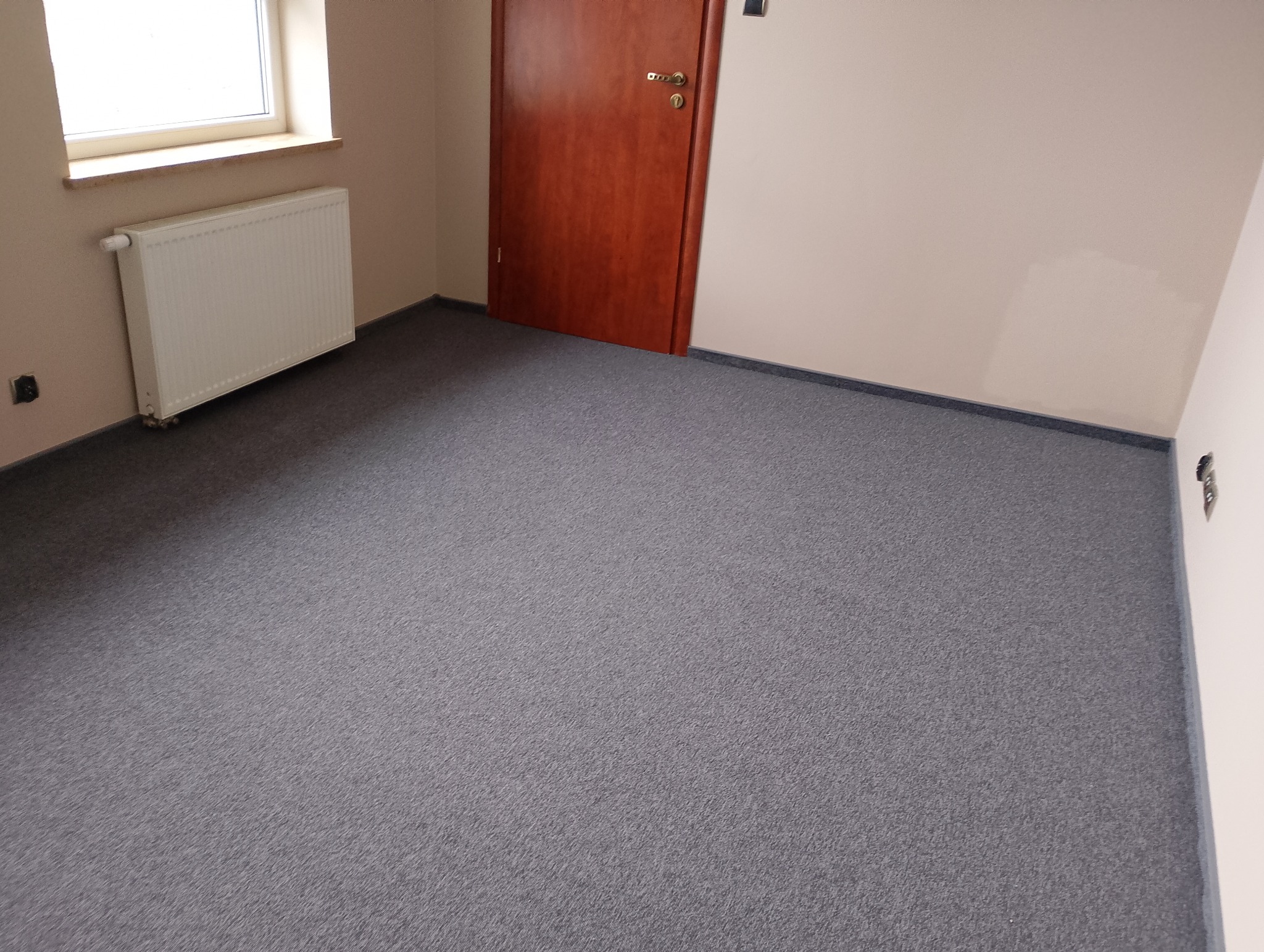 Pusty pokój z szarą wykładziną dywanową, brązowymi drzwiami i białym grzejnikiem pod oknem.