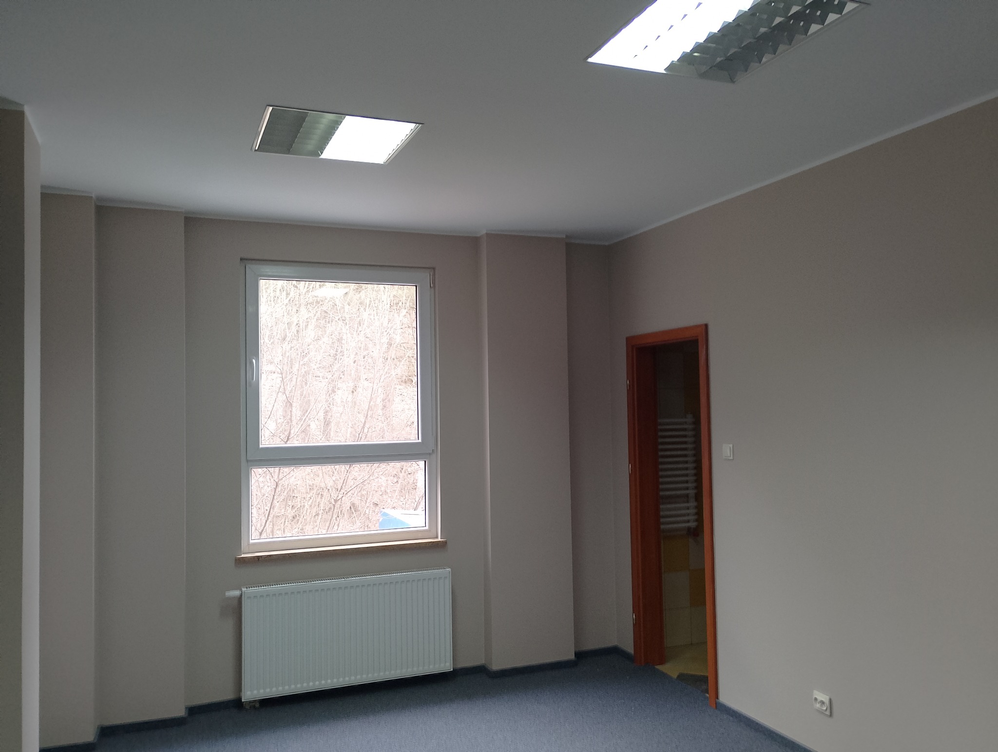 Pomalowane na szaro wnętrze biura z oknem, grzejnikiem i drzwiami do łazienki z widocznym grzejnikiem drabinkowym.