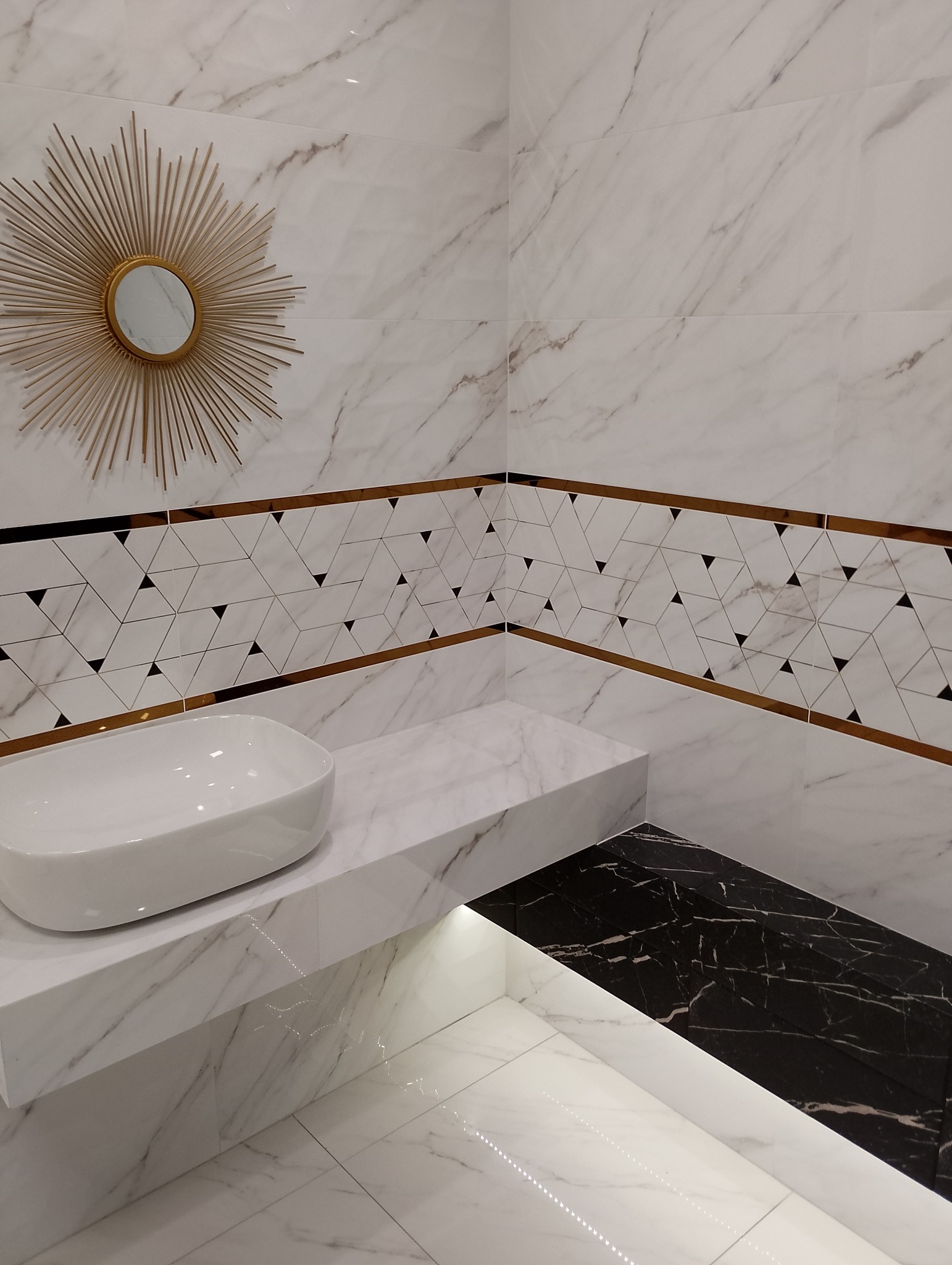 Nowoczesna łazienka z marmurowymi płytkami, umywalką nablatową i lustrem w kształcie słońca. Dekoracyjny pas z geometrycznym wzorem podkreślony złotymi listwami. Podświetlona półka z czarnym marmurem.