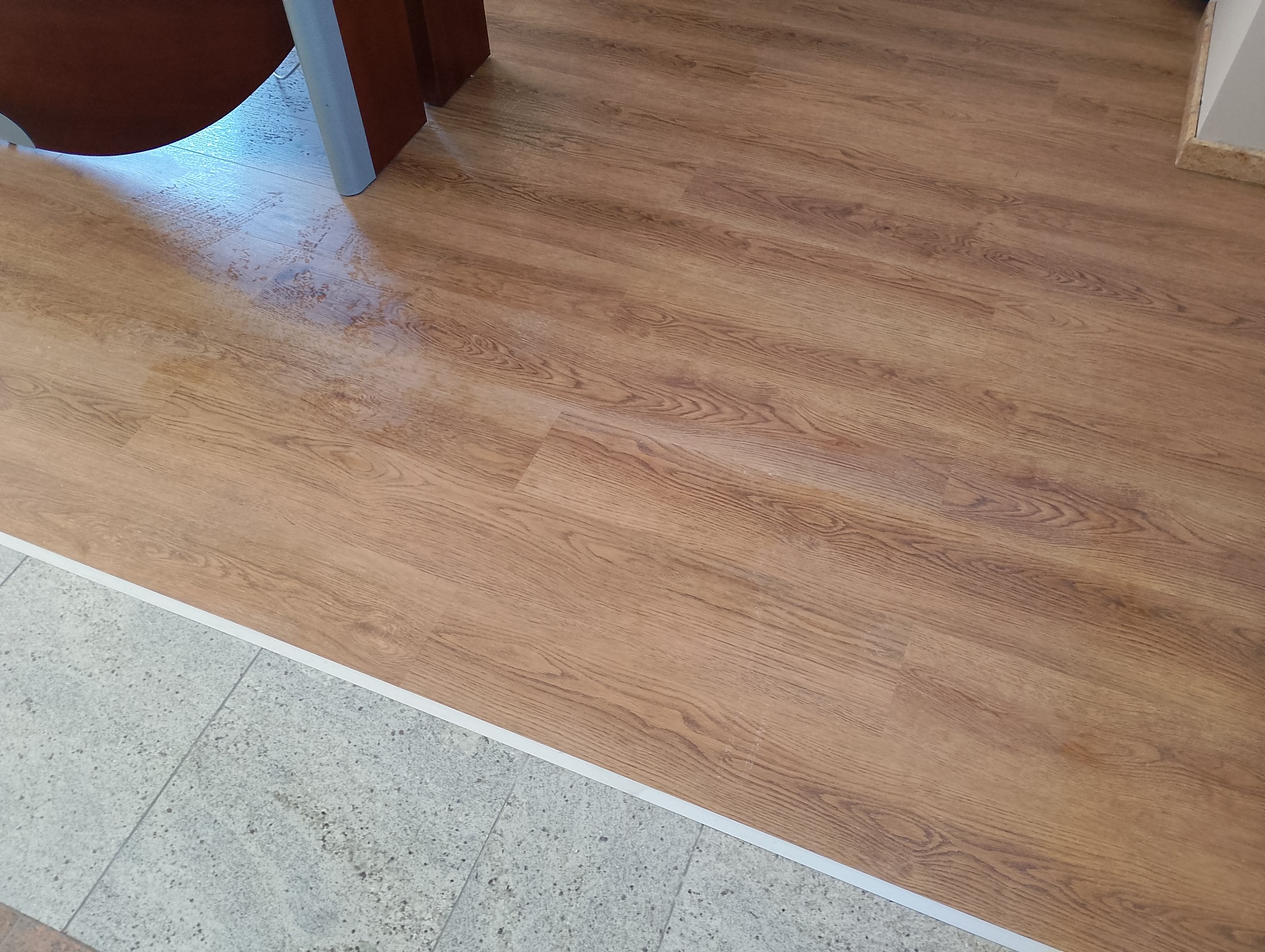 Ułożona podłoga z paneli imitujących drewno w odcieniach brązu, z widocznym przejściem do płytek w odcieniach szarości, wykończonym białą listwą przypodłogową.