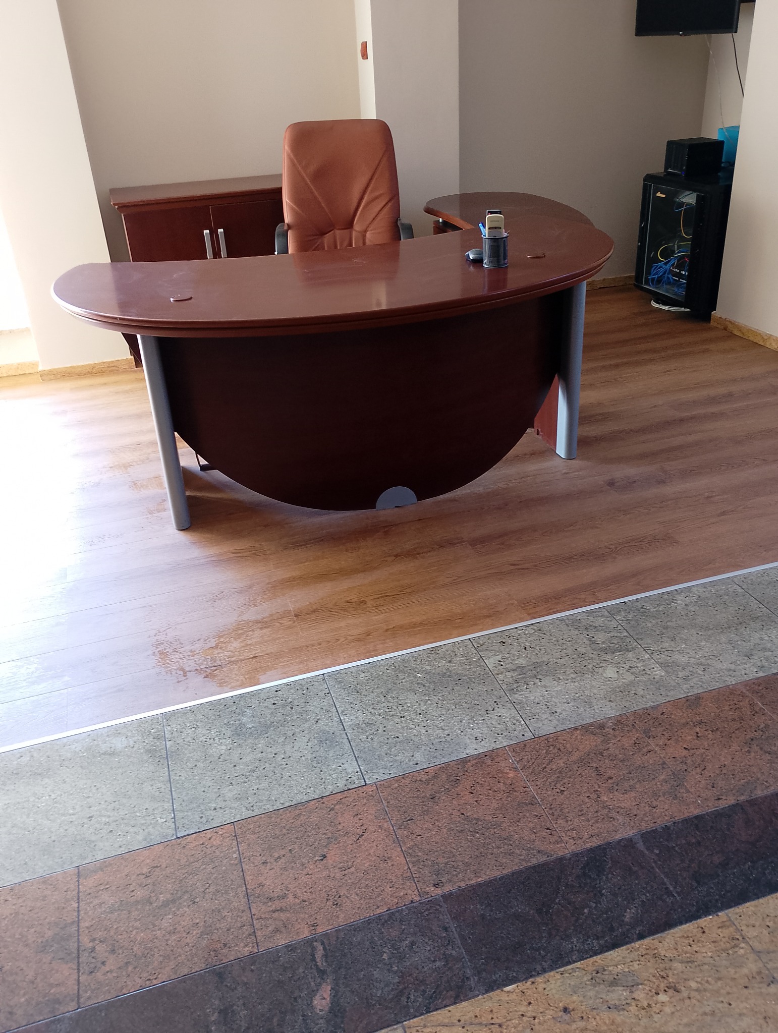 Biuro z brązowym biurkiem o zaokrąglonym kształcie, krzesłem z brązowej skóry, szafką i serwerem w tle, na podłodze panele imitujące drewno i płytki w różnych odcieniach.