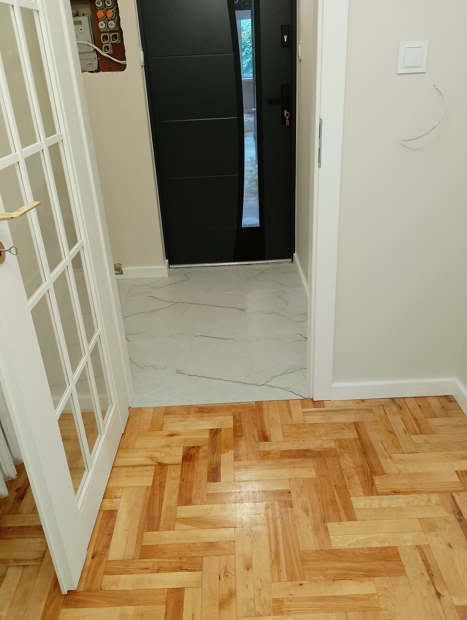 Korytarz po remoncie: parkiet w jodełkę kontrastuje z marmurową posadzką przy czarnych drzwiach wejściowych. Otwarta, biała, przeszklona rama drzwi.