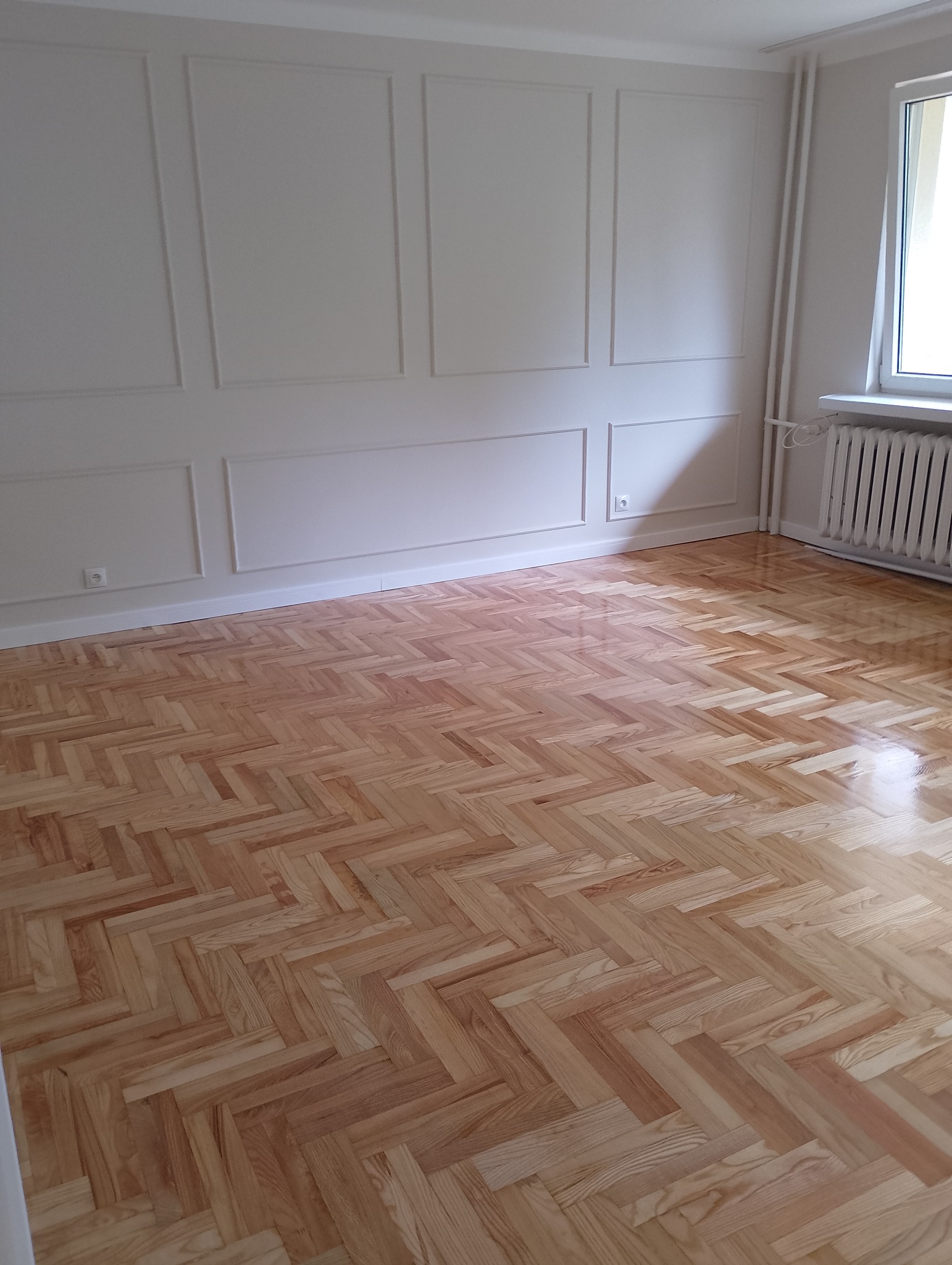 Jasny parkiet ułożony w jodełkę w pustym pokoju z białymi ścianami ozdobionymi sztukaterią. Widoczny grzejnik pod oknem i gniazdko elektryczne.