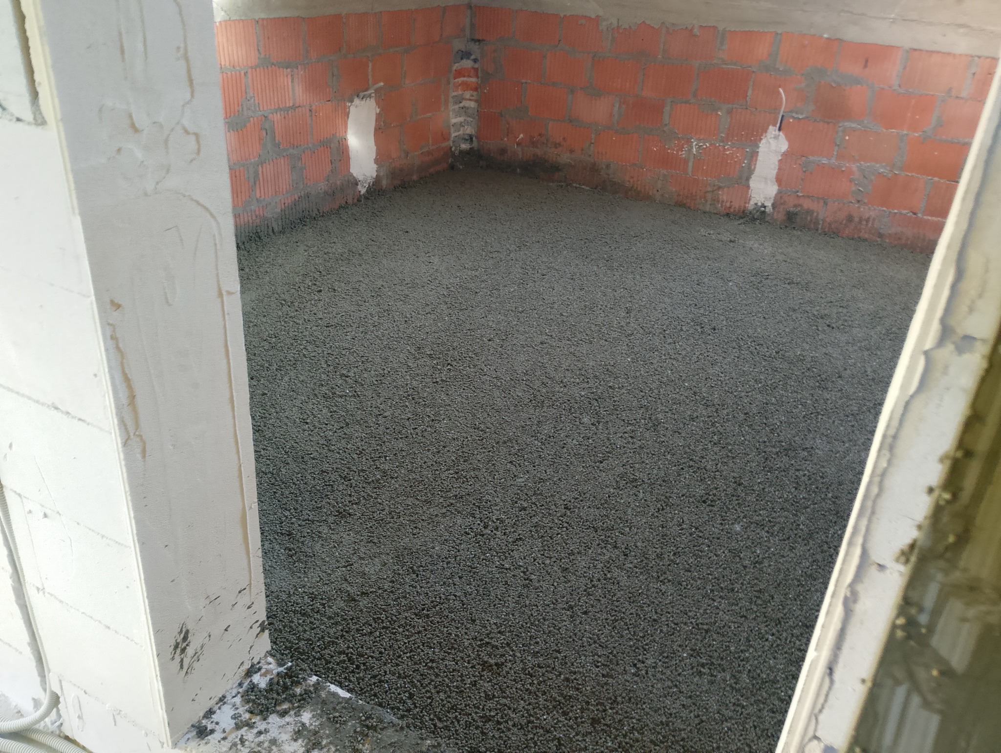 Wylany, świeży podkład podłogowy w pomieszczeniu z surowymi ścianami z czerwonej cegły. Widoczne ślady po instalacjach. Białe, otynkowane futryny drzwi.