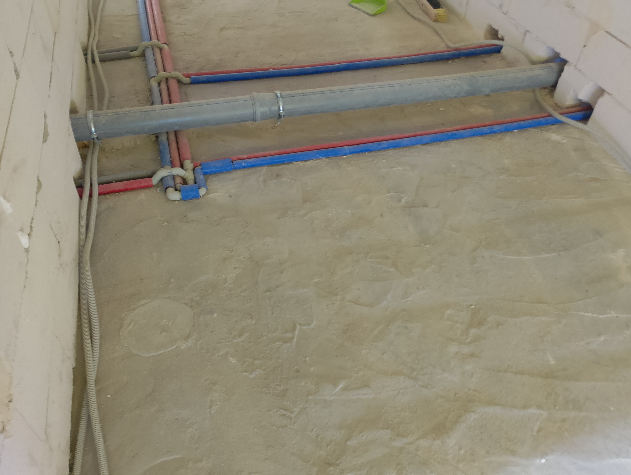 Instalacja wodno-kanalizacyjna i elektryczna w bruzdach pod wylewką. Widoczne rury PEX w kolorze niebieskim i czerwonym oraz peszle z kablami. Ściany z betonu komórkowego.