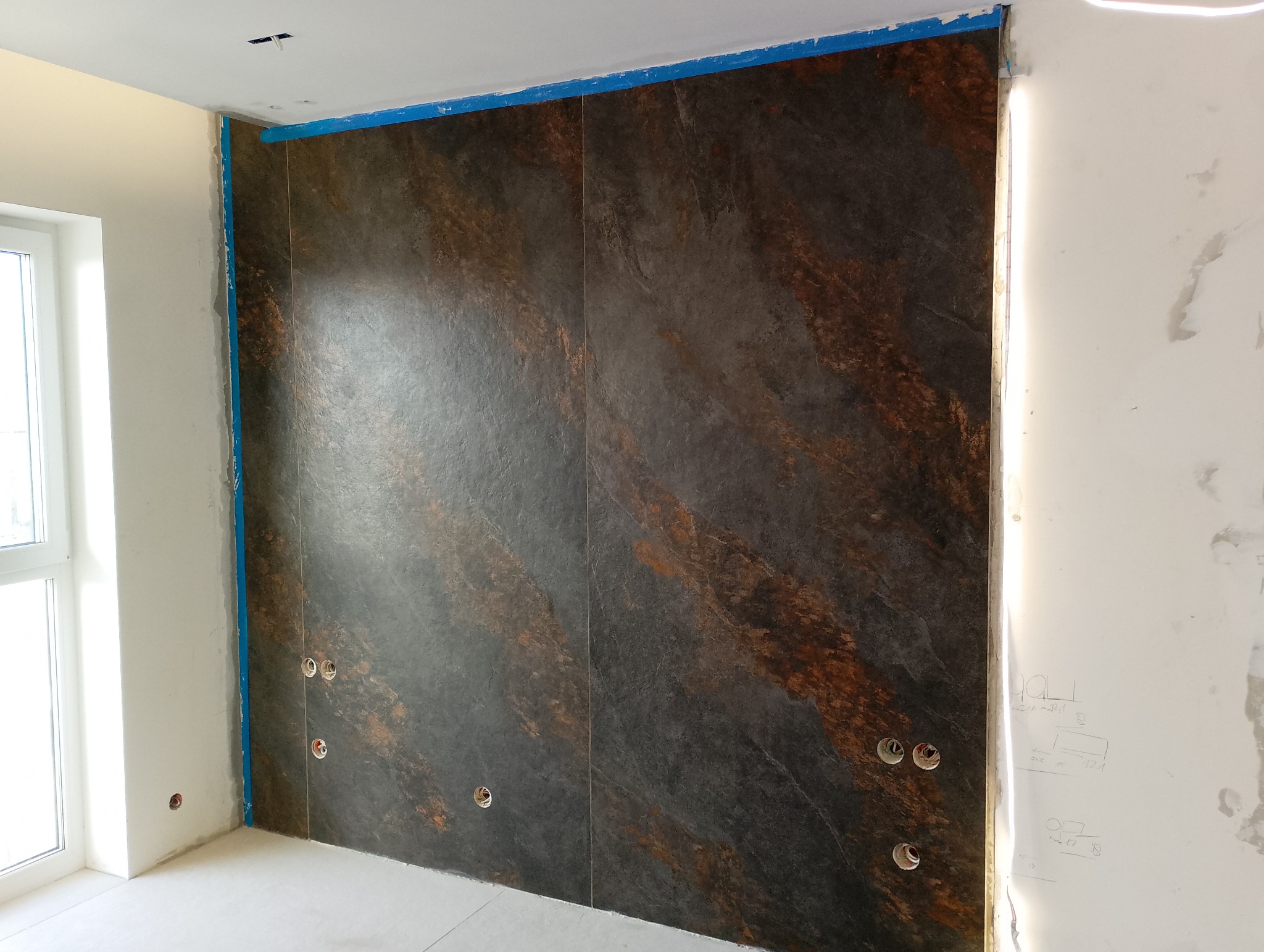 Ściana wyłożona ciemnymi płytami imitującymi kamień z widocznymi otworami na gniazdka elektryczne, zabezpieczona niebieską taśmą malarską przy suficie i ścianach.